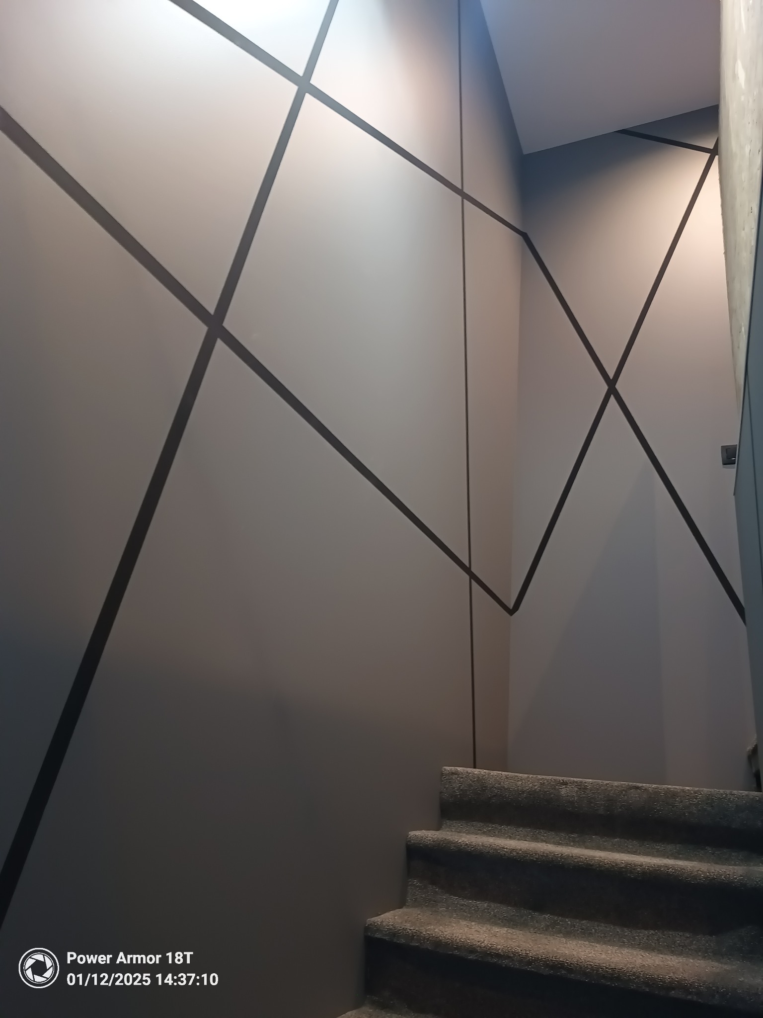 Ściana klatki schodowej z geometrycznym wzorem z ciemnych pasów, szare schody pokryte wykładziną dywanową.