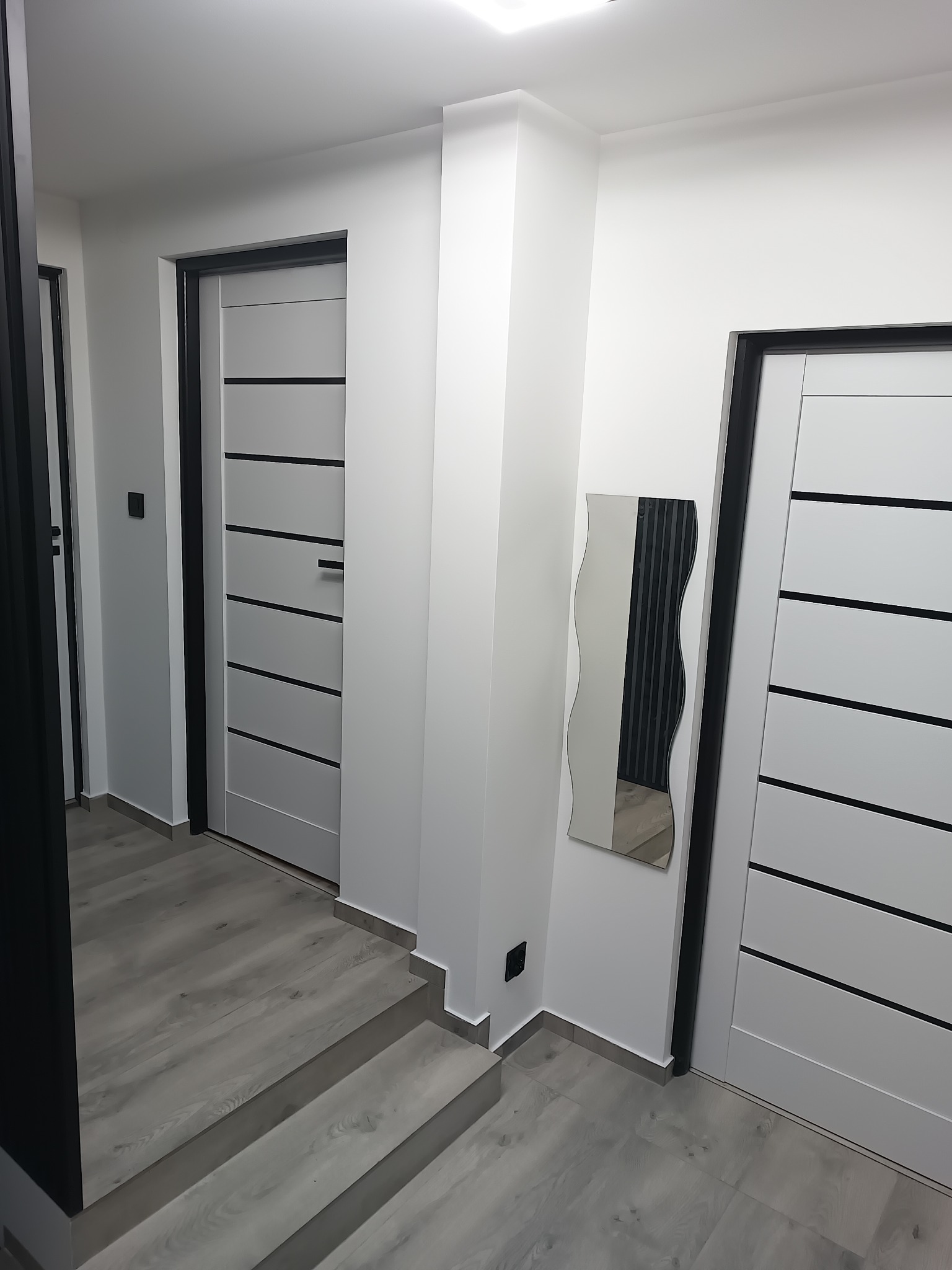 Nowoczesny przedpokój w odcieniach szarości i bieli z trzema drzwiami z czarnymi ościeżnicami i poziomymi paskami, ozdobiony falistym lustrem i panelami podłogowymi imitującymi drewno.