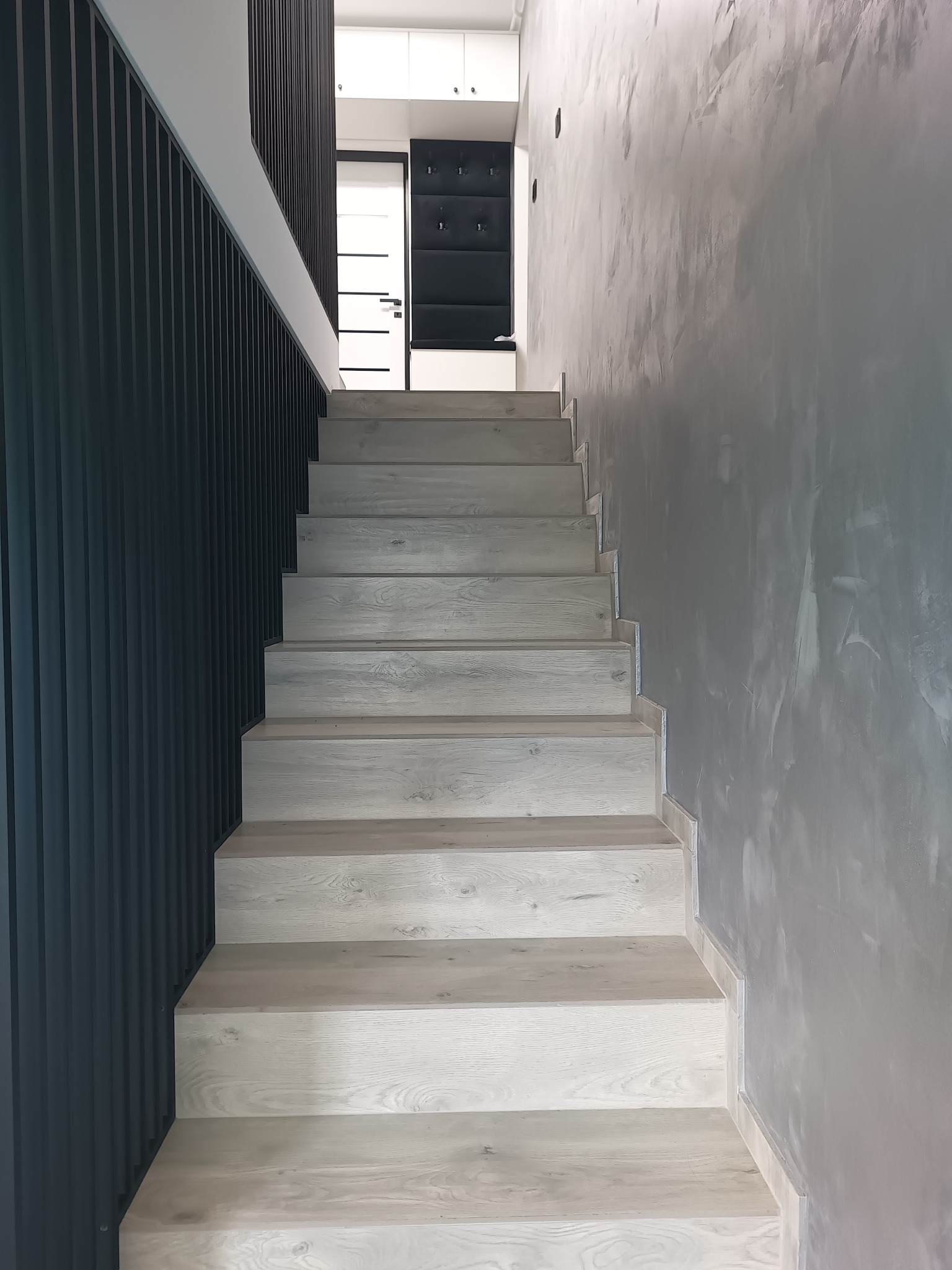 Schody z jasnego drewna prowadzące do góry, obok ściana z ciemnymi pionowymi panelami i ściana z szarą gładzią dekoracyjną.
