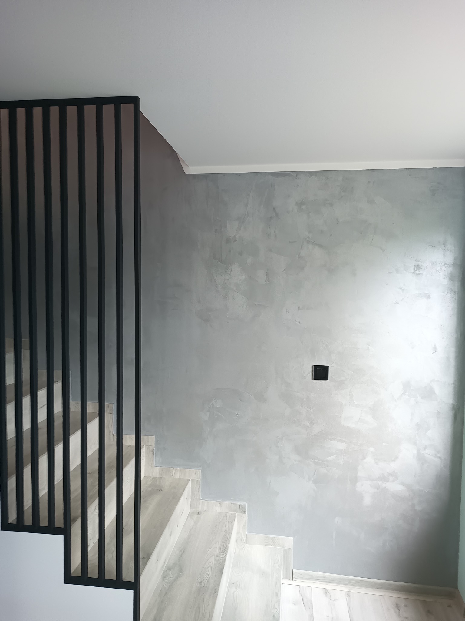 Ściana z dekoracyjną szarą strukturą w Jaroszowie, widoczna obok schodów z czarną, metalową balustradą i gniazdkiem elektrycznym w ścianie.