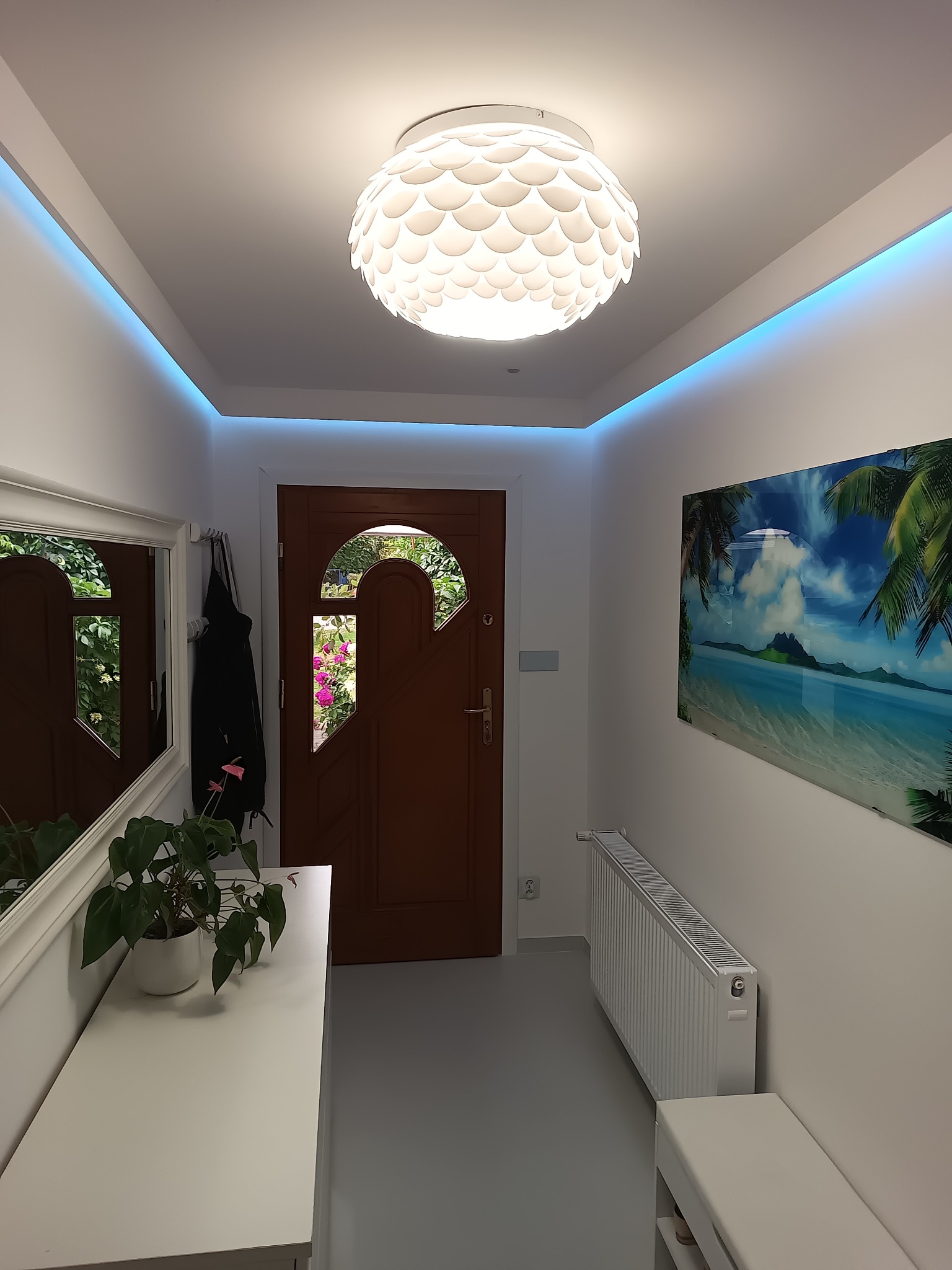 Nowoczesny przedpokój z białymi ścianami, podświetlanym sufitem, drewnianymi drzwiami wejściowymi z dekoracyjnym przeszkleniem, lustrem w białej ramie, komodą z rośliną doniczkową i obrazem...