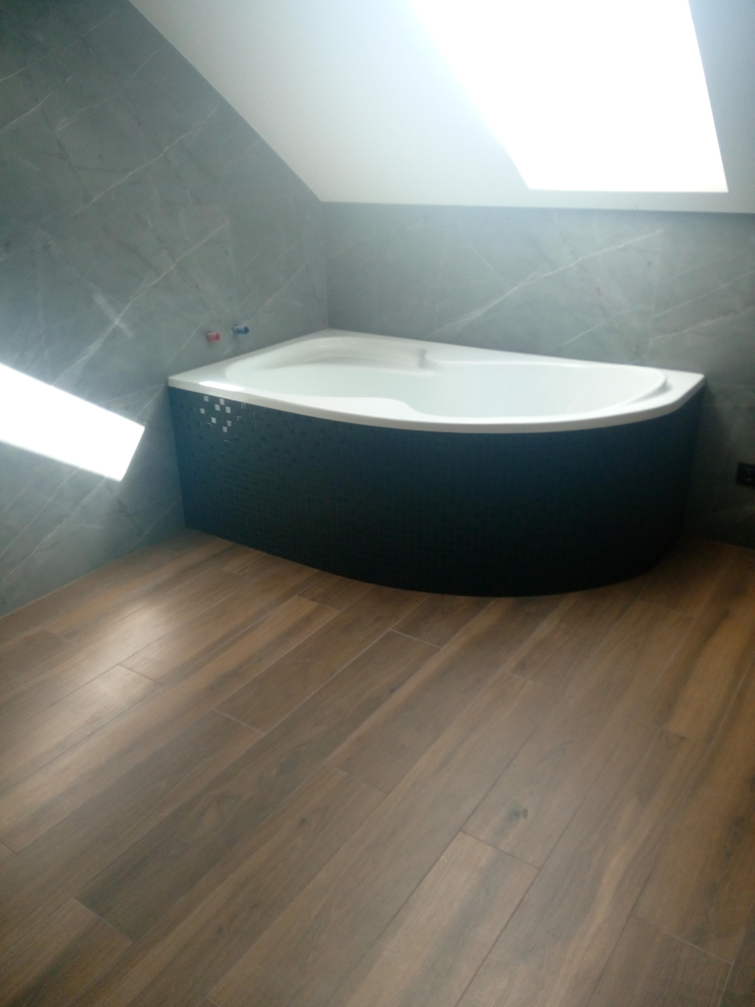 Narożna wanna z czarną mozaiką na obudowie, szare płytki imitujące marmur na ścianach i podłoga wyłożona panelami imitującymi drewno w łazience ze skośnym sufitem i oknem dachowym.