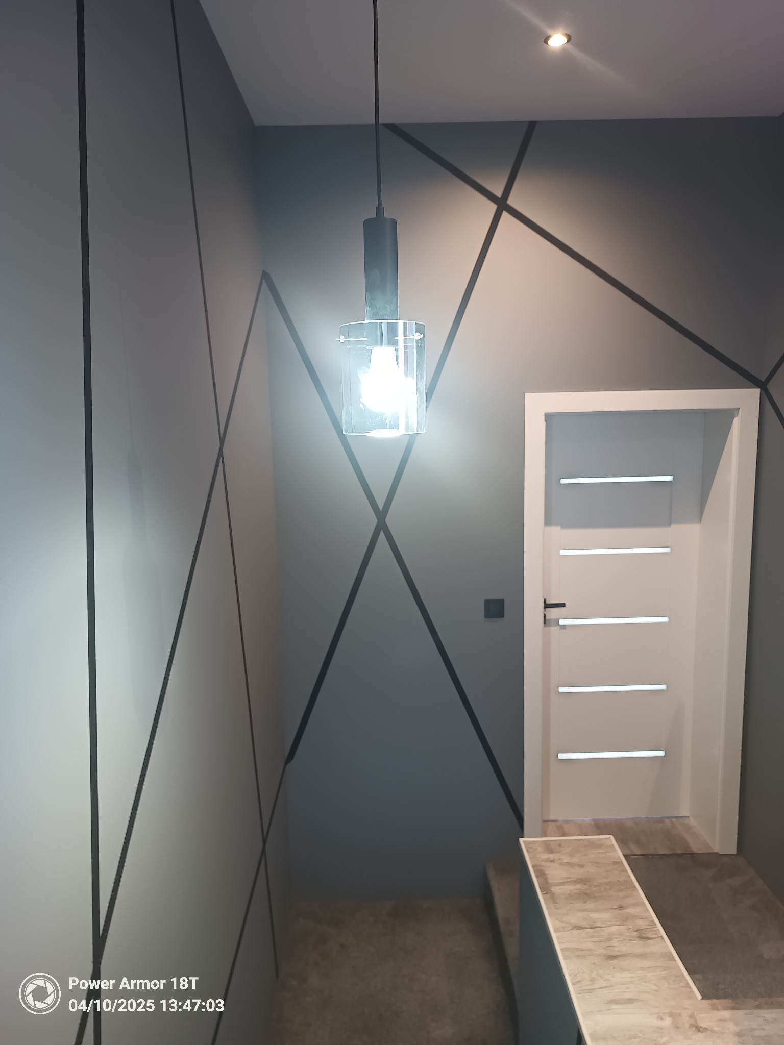 Nowoczesne wykończenie wnętrza: szare ściany z geometrycznym wzorem z czarnych linii, białe drzwi z poziomymi przeszkleniami i wisząca lampa z przezroczystym kloszem.