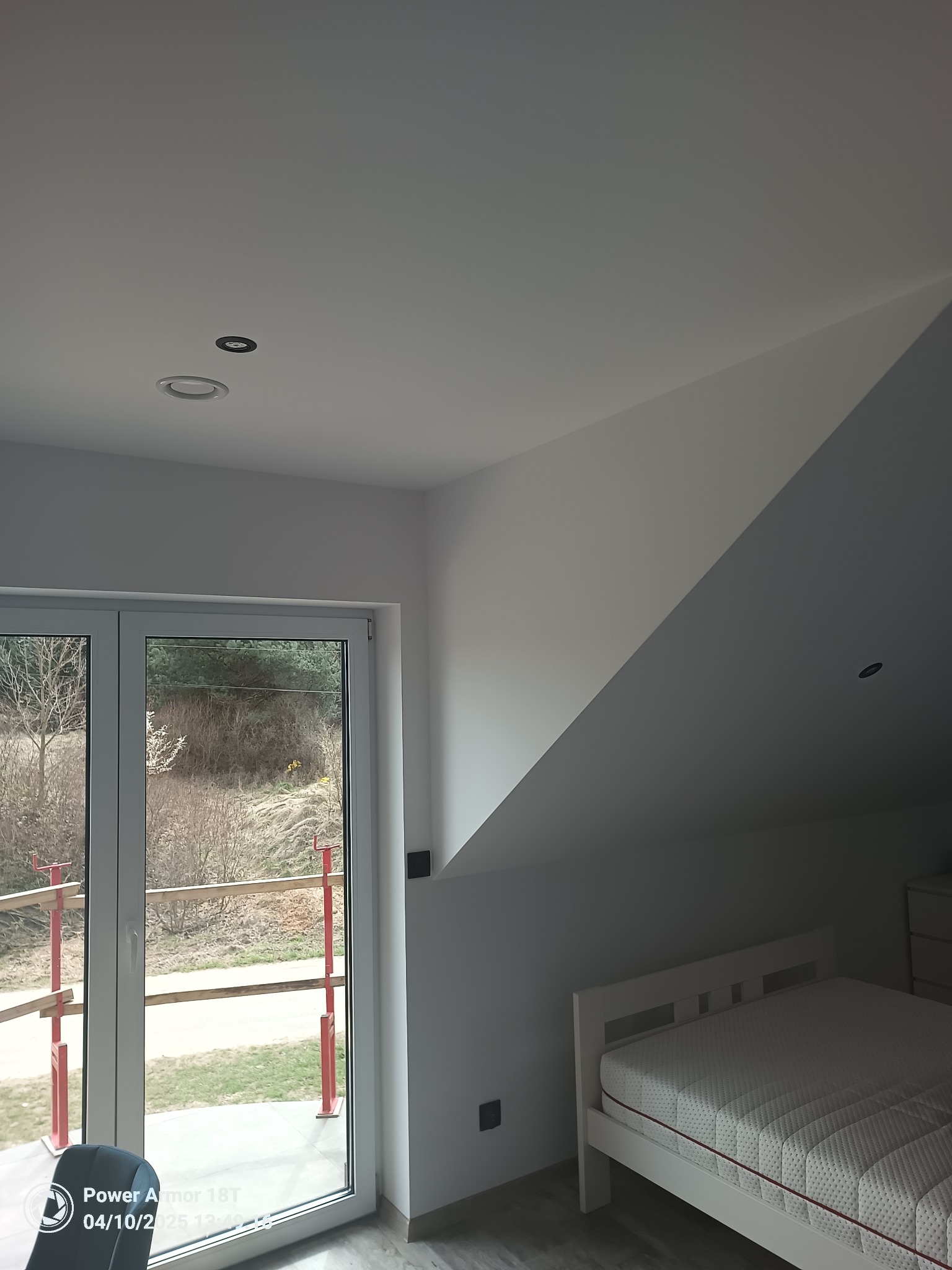 Poddasze po remoncie: białe ściany, skosy, okno balkonowe z widokiem na zewnątrz, białe łóżko z materacem i szara podłoga.