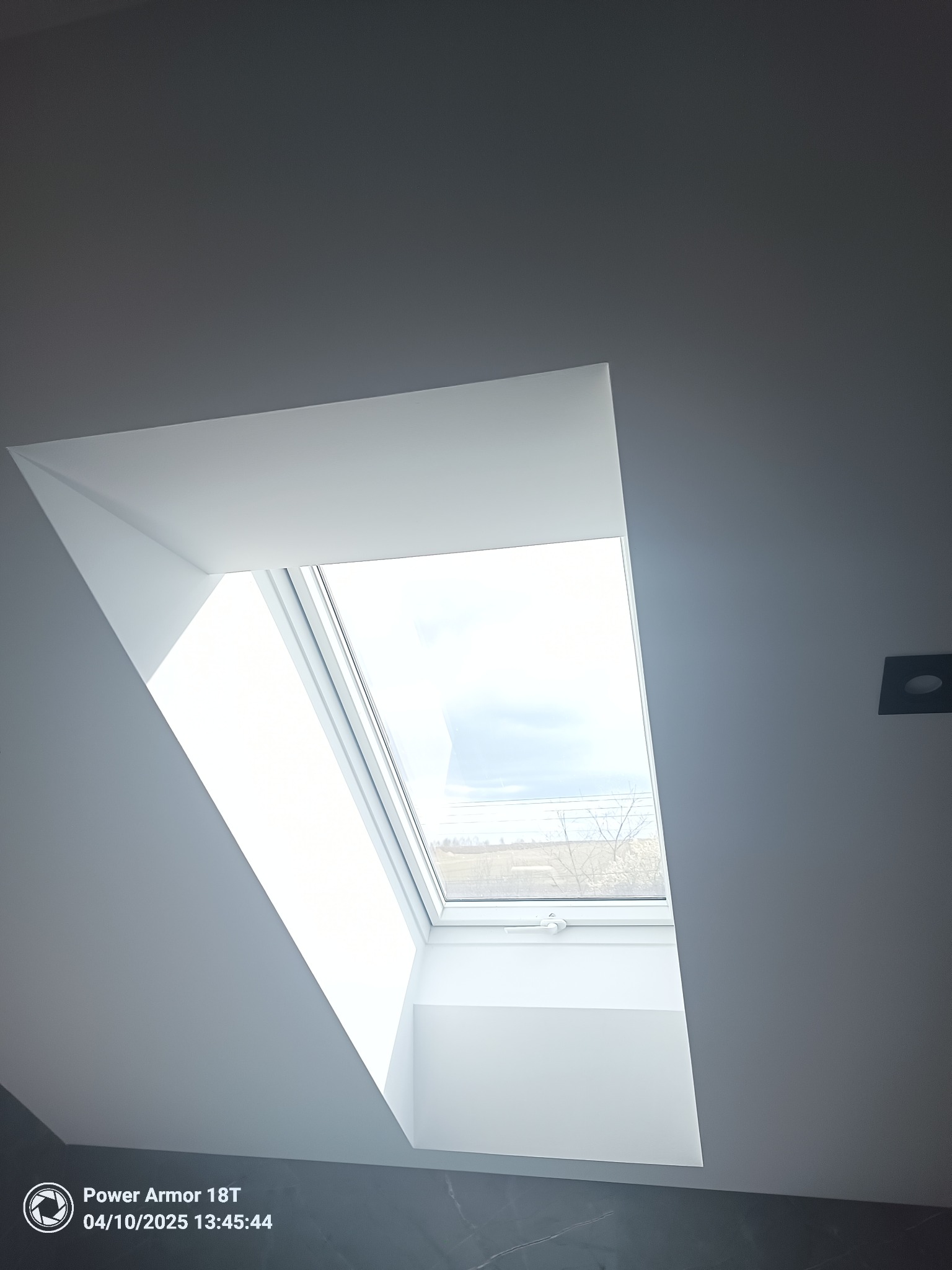 Wnętrze poddasza z oknem dachowym, widok na zewnątrz z drzewami, szare ściany i marmurowe płytki podłogowe.