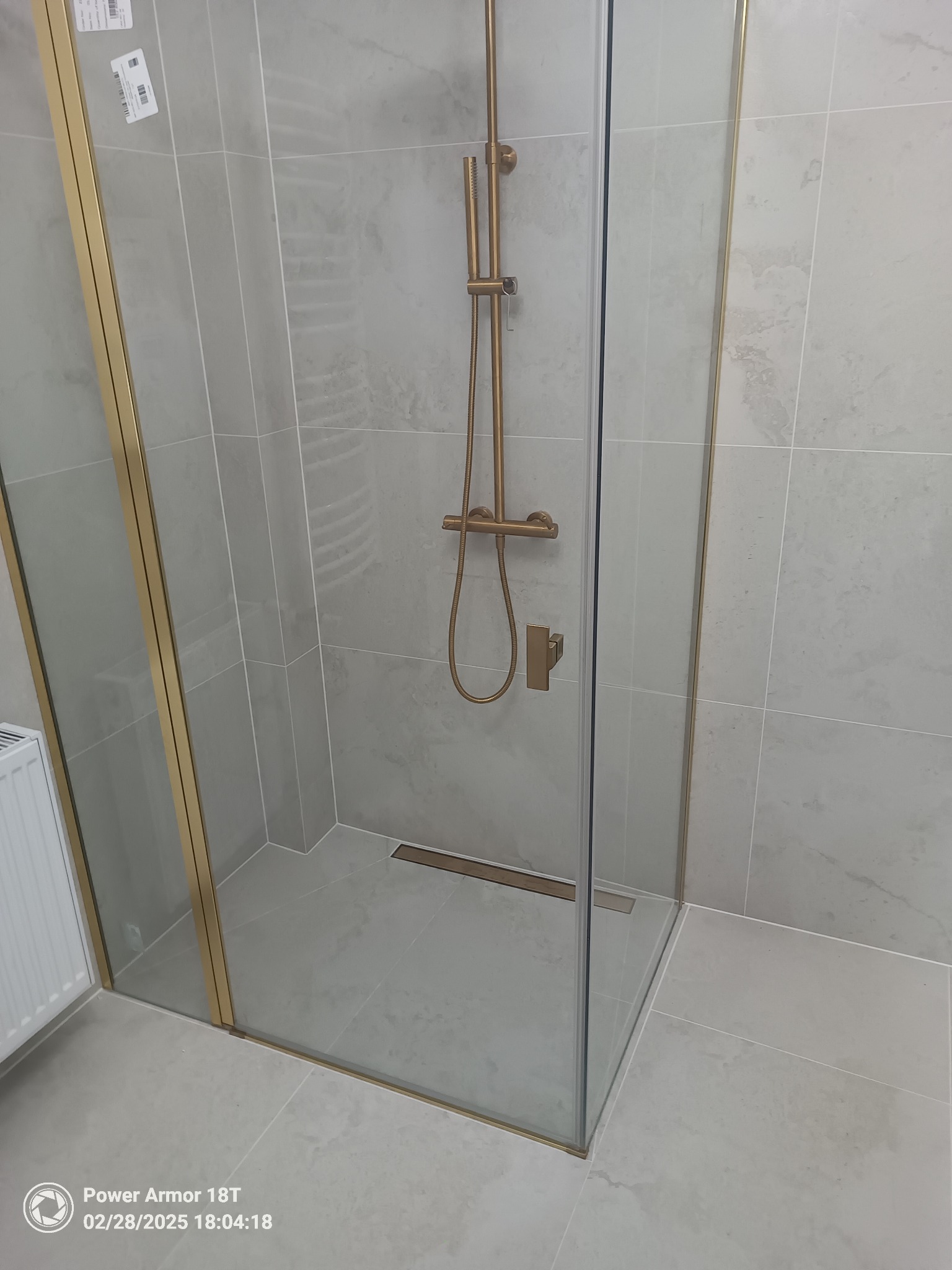 Nowoczesna kabina prysznicowa z szklanymi drzwiami i złotymi profilami, szare płytki na ścianach i podłodze, odpływ liniowy w podłodze.