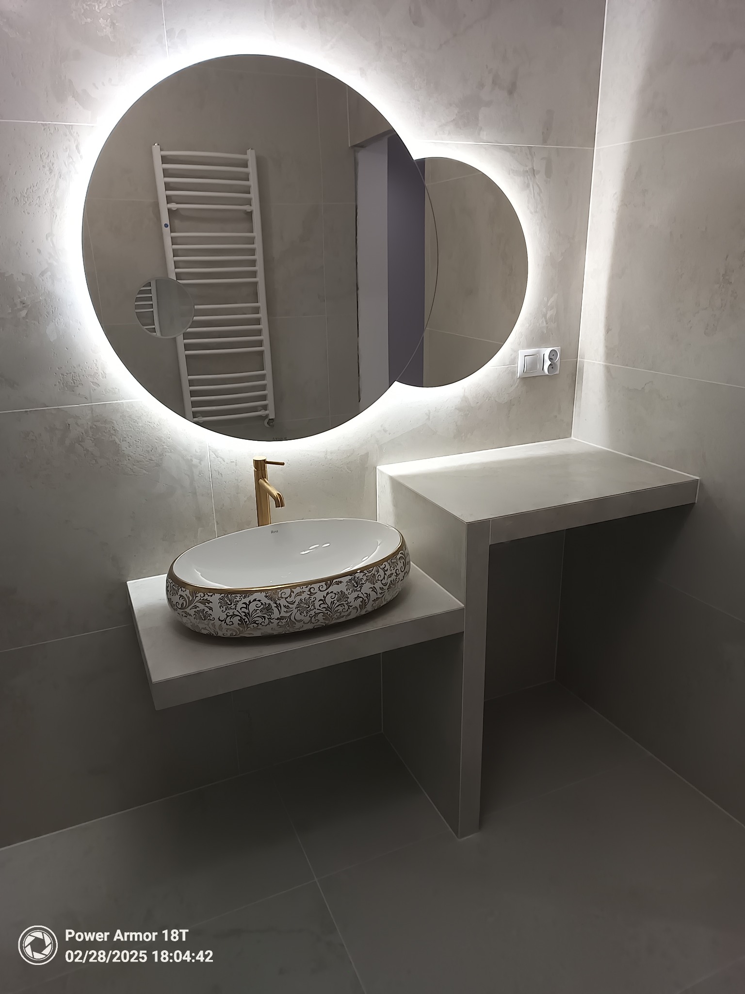 Nowoczesna łazienka z umywalką nablatową w kolorze białym z dekoracyjnym złotym wzorem, złotą baterią i okrągłym lustrem z podświetleniem LED, grzejnik drabinkowy widoczny w odbiciu lustra.