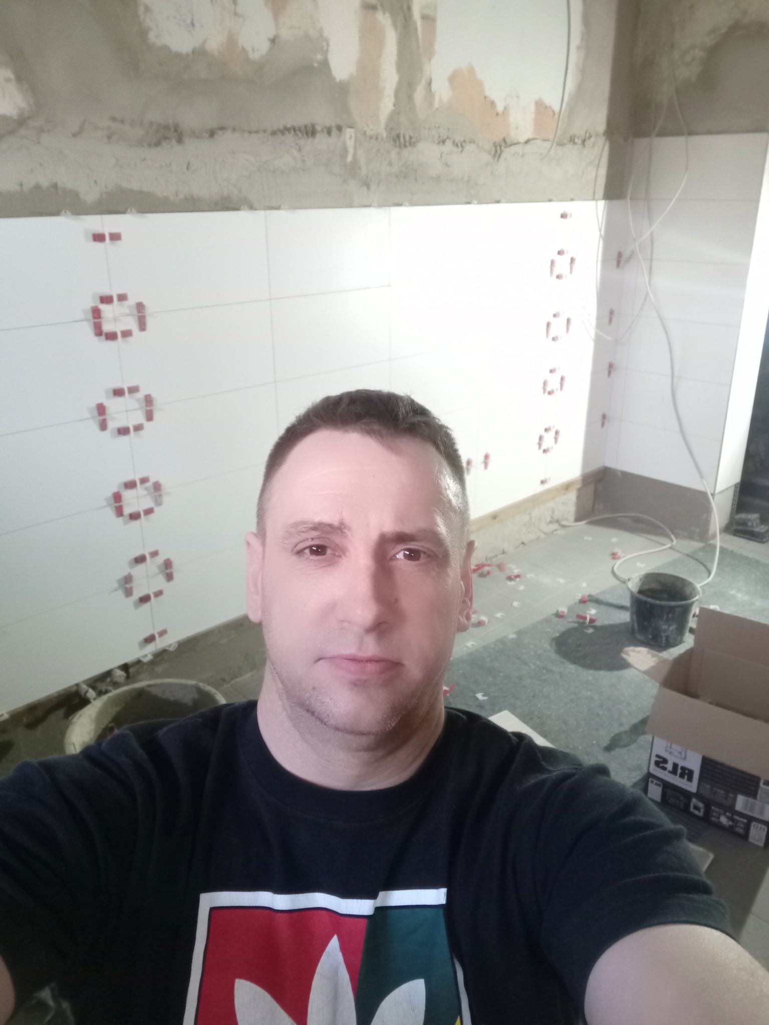 Mężczyzna robi selfie na tle ściany z częściowo ułożonymi białymi płytkami, z widocznymi krzyżykami dystansowymi i surowym wykończeniem ściany nad płytkami, na podłodze resztki zaprawy i krzyżyki.