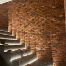 Ściana wykończona czerwoną cegłą klinkierową przylegająca do schodów z podświetleniem LED, widoczne niedociągnięcia w fugowaniu cegły.