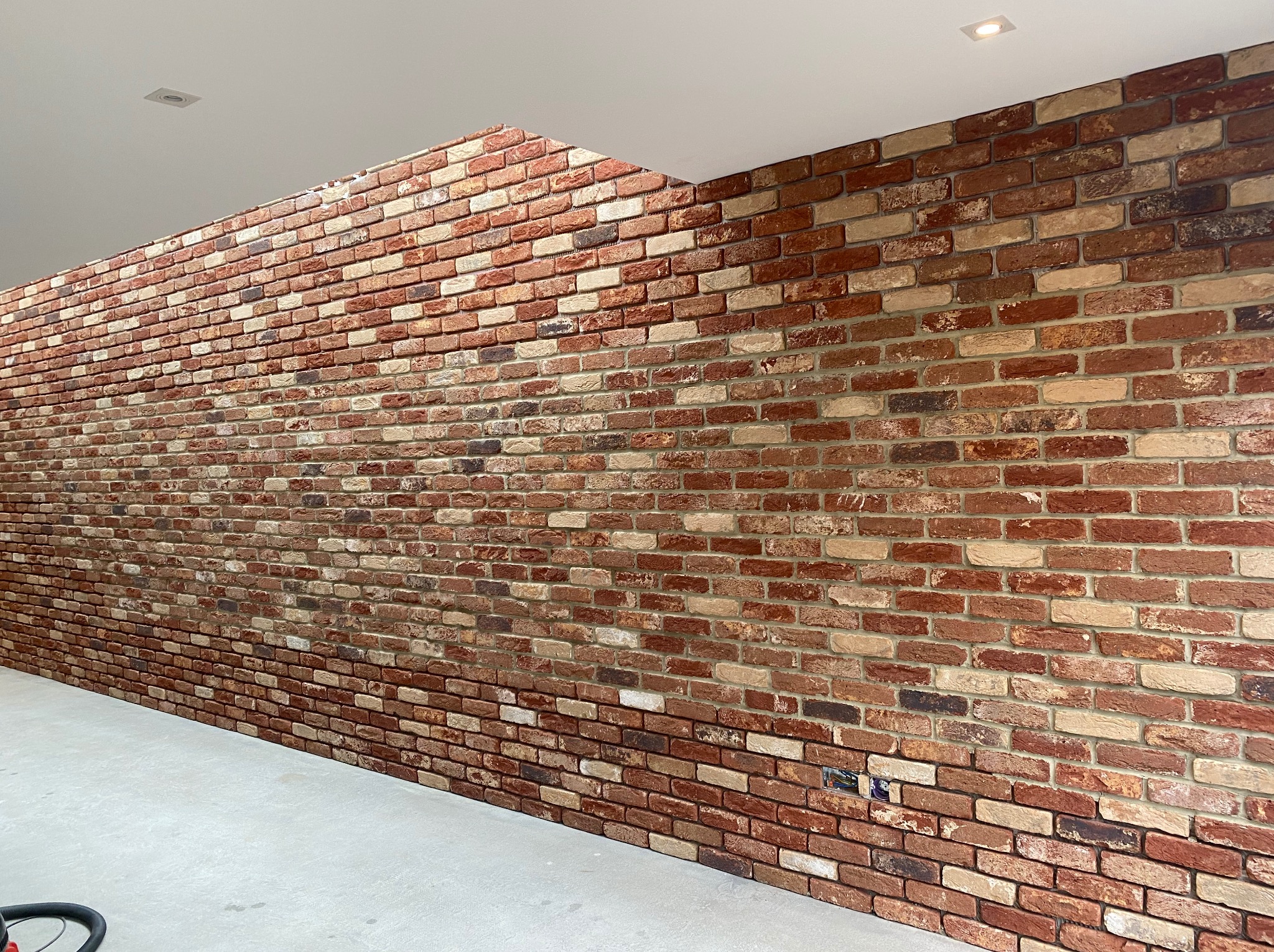Ściana z czerwonej cegły wewnątrz pomieszczenia, z widocznym oświetleniem sufitowym i szarą podłogą, ukazująca detale faktury i kolorystyki cegieł.