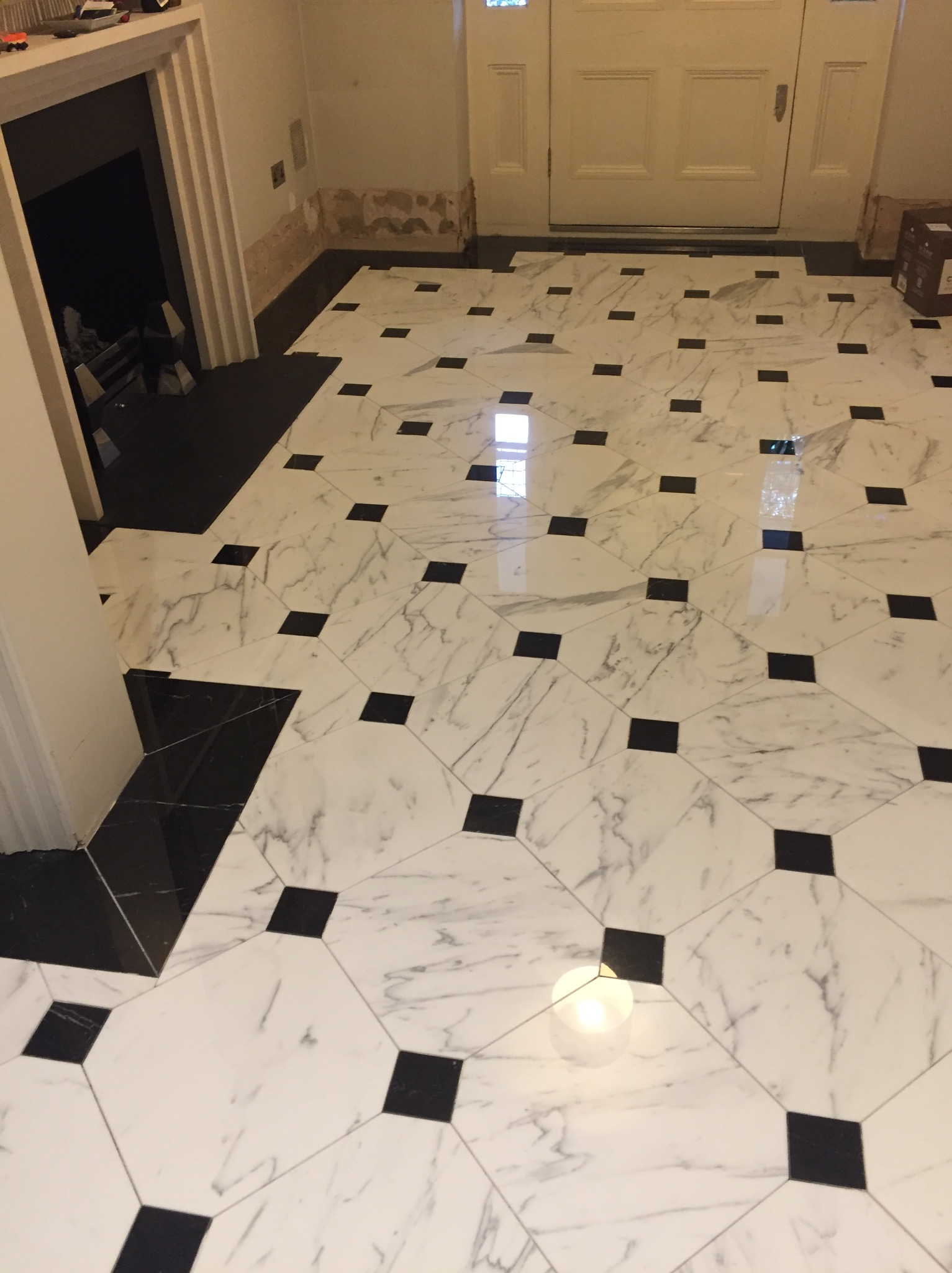 Wykończona podłoga z białych płytek imitujących marmur z czarnymi kwadratami, widoczny fragment ściany z uszkodzonym tynkiem i białe drzwi wejściowe.