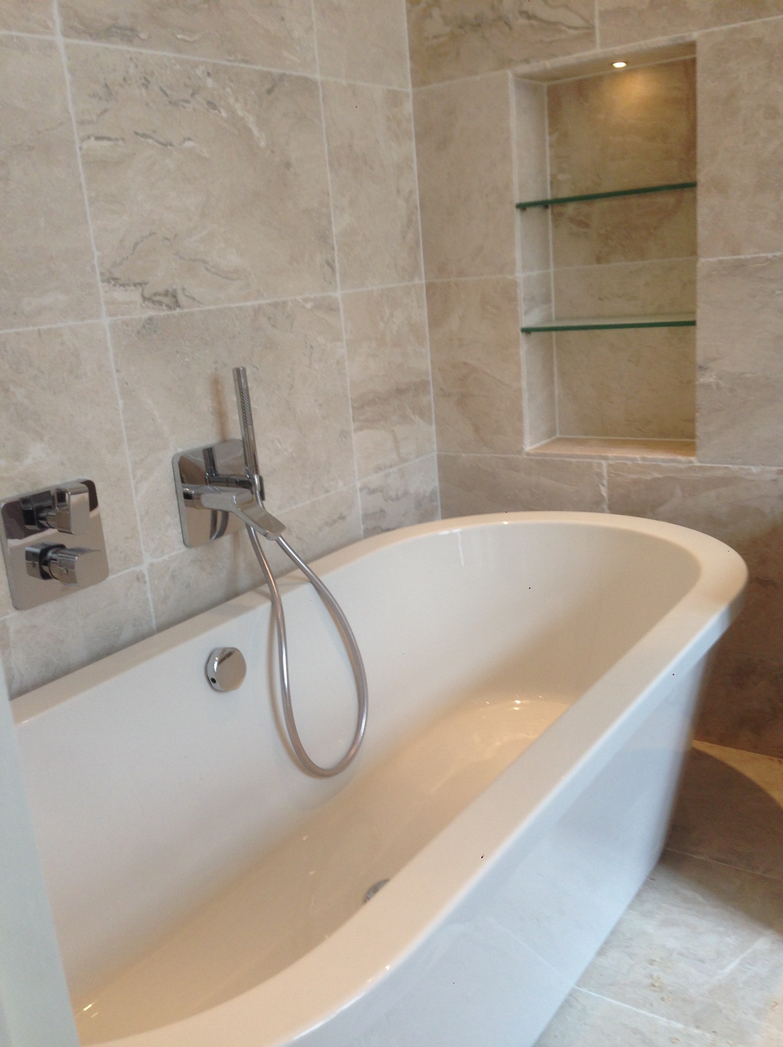 Biała wanna wolnostojąca z chromowaną armaturą i wnęką z szklanymi półkami w ścianie wyłożonej beżowymi płytkami imitującymi marmur.