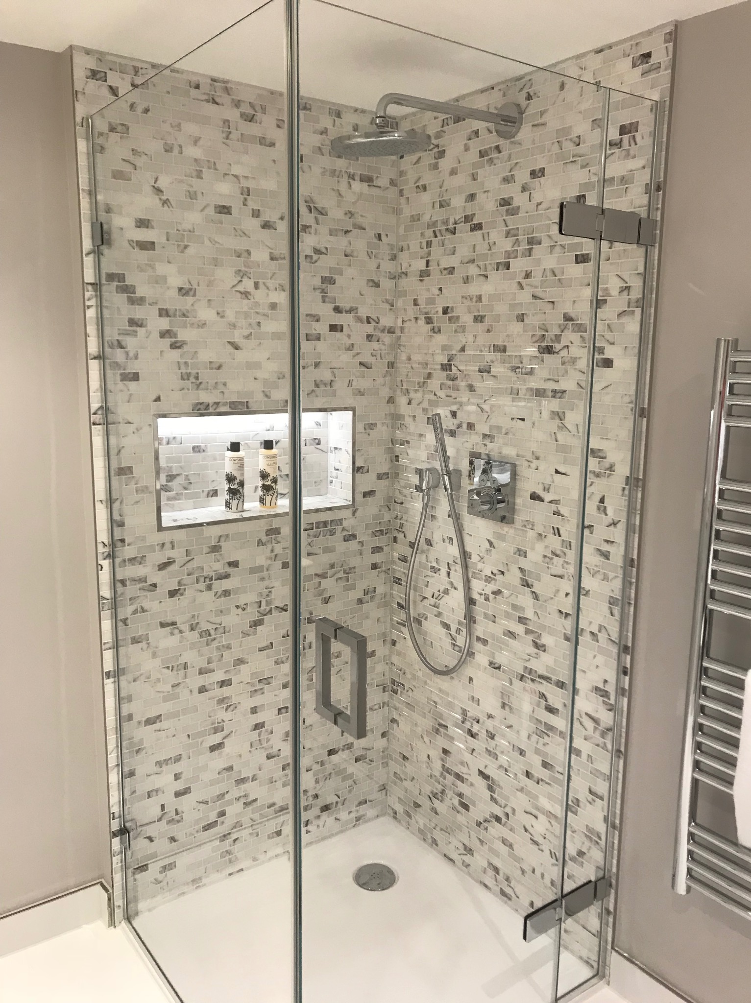 Szklana kabina prysznicowa wykończona mozaiką w odcieniach szarości i bieli, z wnęką na kosmetyki, chromowaną armaturą i grzejnikiem drabinkowym z ręcznikiem.