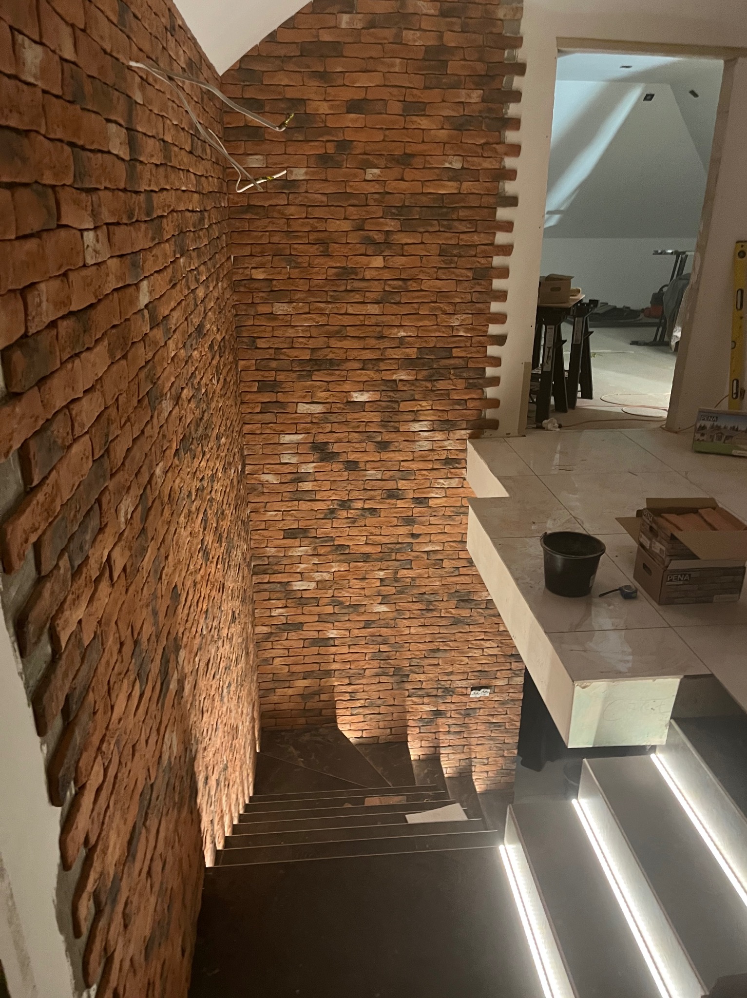 Korytarz w trakcie wykańczania z ceglaną ścianą, podświetlanymi schodami i widocznymi przewodami elektrycznymi.