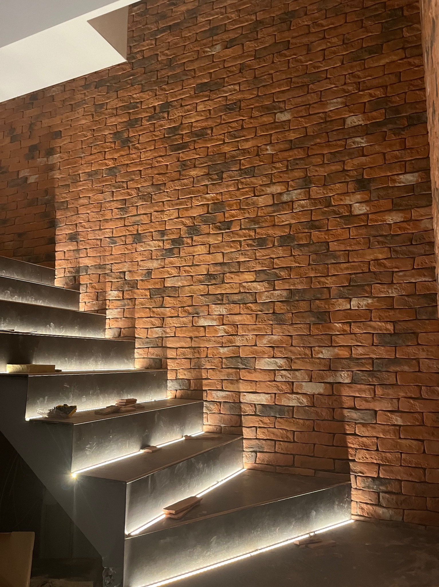 Ściana wykończona czerwoną cegłą klinkierową przylegająca do schodów z podświetleniem LED, widoczne niedociągnięcia w fugowaniu cegły.