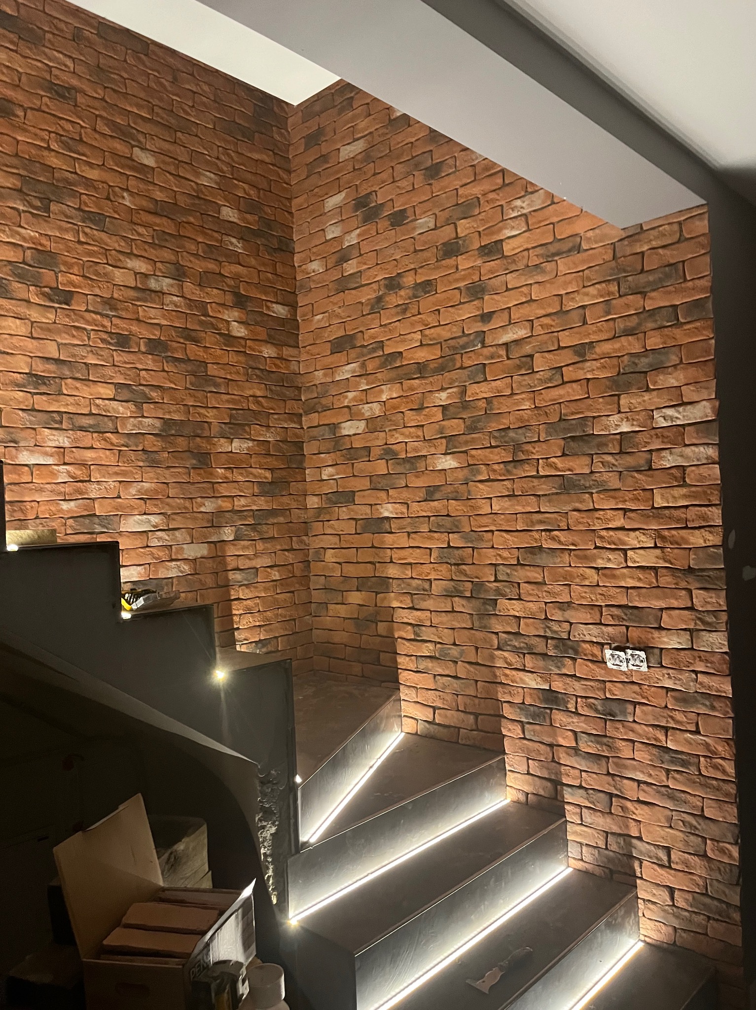 Ściana z cegły klinkierowej w korytarzu ze schodami podświetlonymi LED, widoczne puszki elektryczne i fragment kartonu z materiałami budowlanymi.