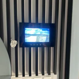 Czarny wideodomofon marki Vidos zamontowany na ścianie z dekoracyjnymi, pionowymi listwami w kolorze czarno-białym, wyświetlający obraz z kamery zewnętrznej. Widoczne przyciski funkcyjne na panelu...