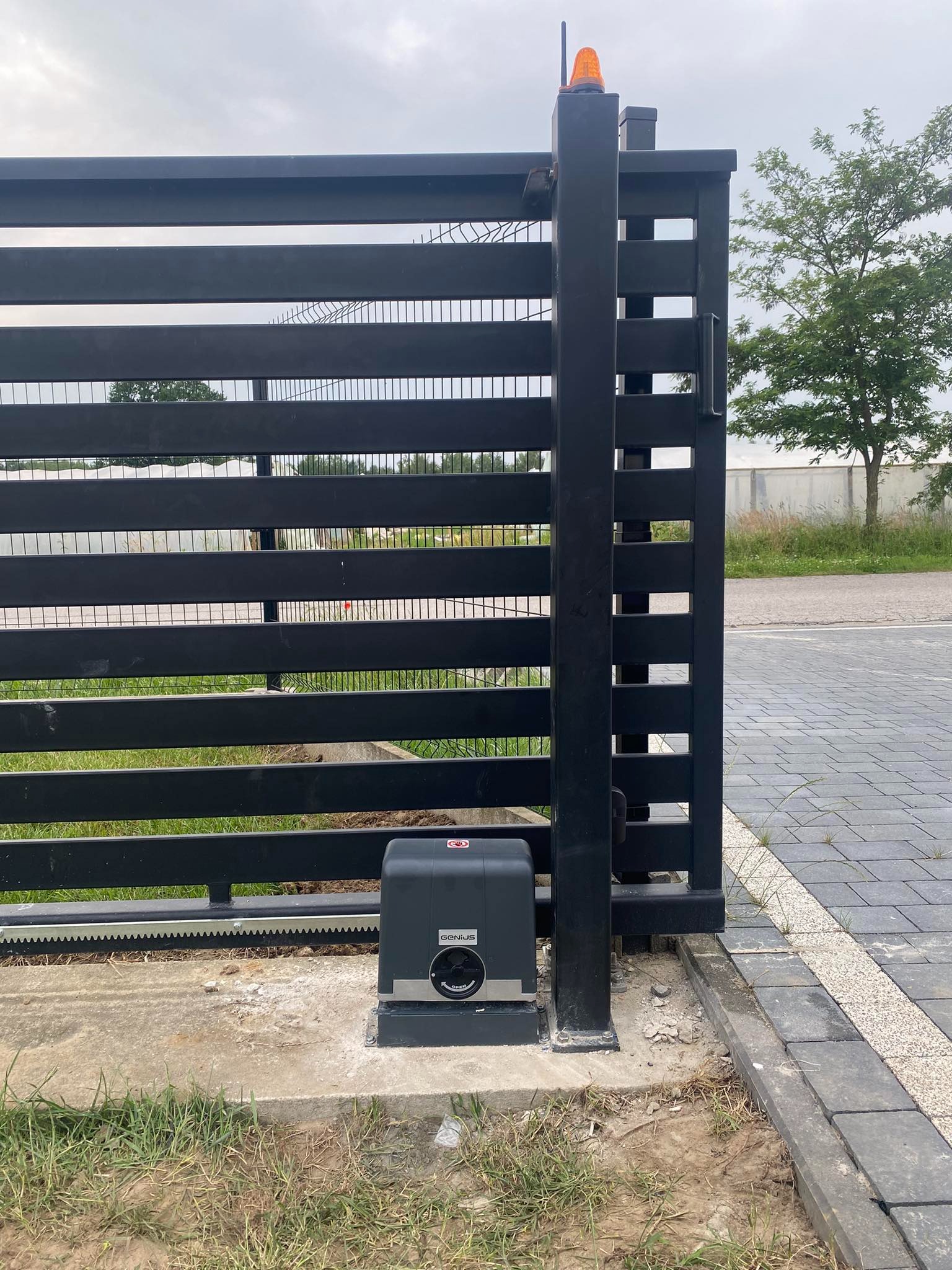 Czarna, metalowa brama przesuwna z zamontowanym napędem Genius na betonowej podstawie, widoczny sygnalizator świetlny, kostka brukowa i trawa.