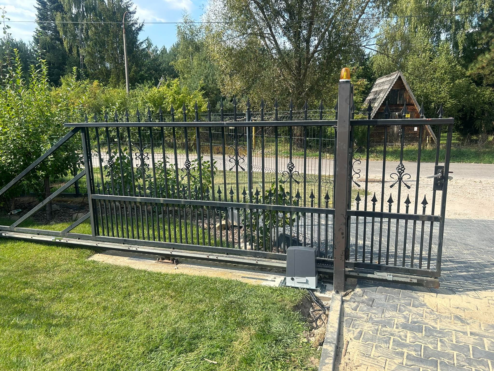 Czarna, przesuwna brama wjazdowa z automatyką, widoczny napęd i listwa zębata, trawnik i kostka brukowa, w tle zieleń i mały drewniany budynek.