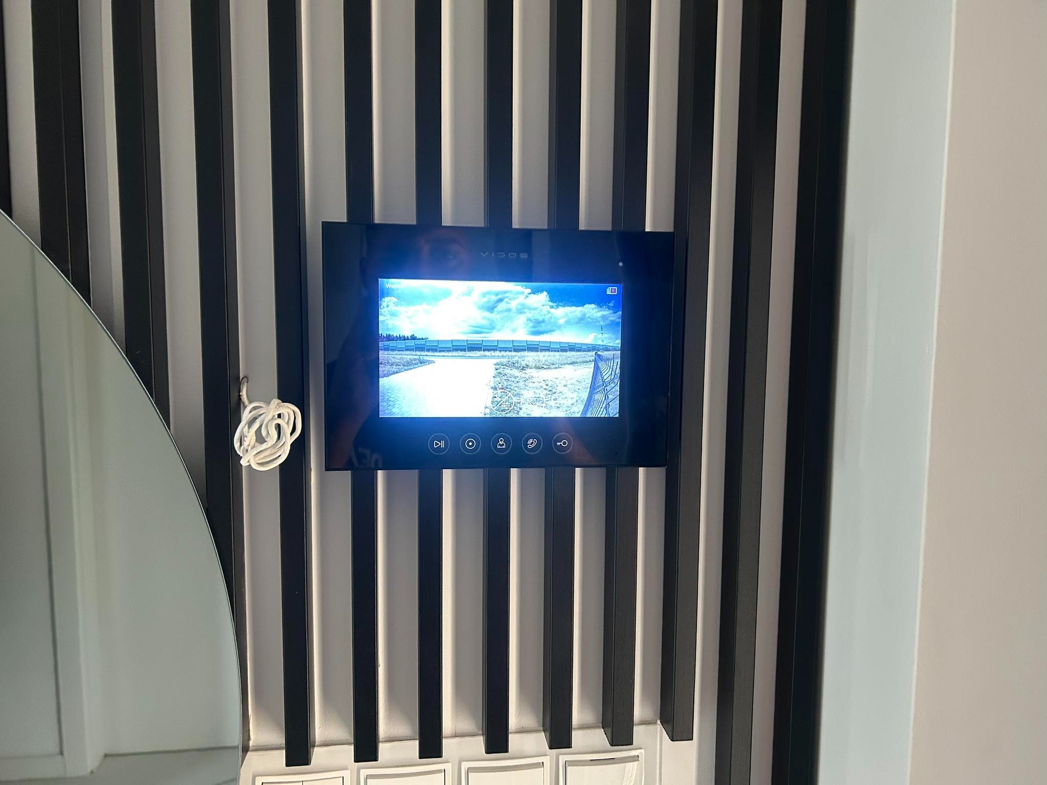 Czarny wideodomofon marki Vidos zamontowany na ścianie z dekoracyjnymi, pionowymi listwami w kolorze czarno-białym, wyświetlający obraz z kamery zewnętrznej. Widoczne przyciski funkcyjne na panelu...