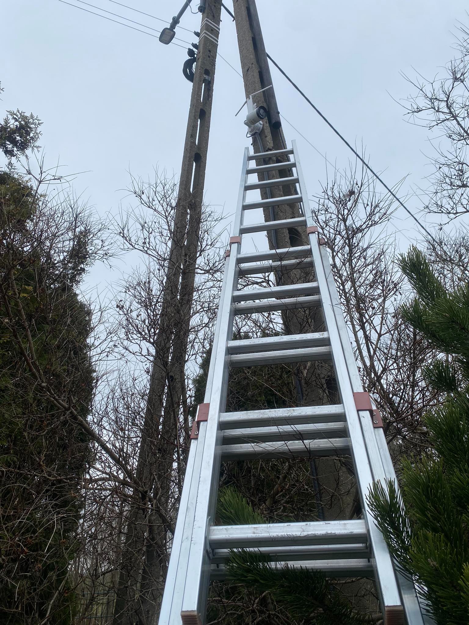 Aluminiowa drabina oparta o betonowy słup energetyczny z zamontowanym reflektorem i kamerą CCTV, widok z dołu na tle pochmurnego nieba i drzew.