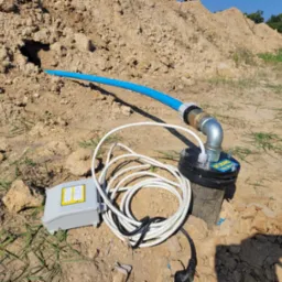 Instalacja studni na piaszczystym terenie: niebieska rura wystaje z wykopu, podłączona do metalowego złącza i czarnej obudowy z naklejką, obok szare pudełko sterujące i zwinięty kabel zasilający.
