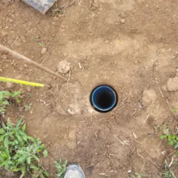 Wykopany otwór w ziemi z niebieską rurą w środku, obok widoczne narzędzia pomiarowe, buty robocze i roślinność.
