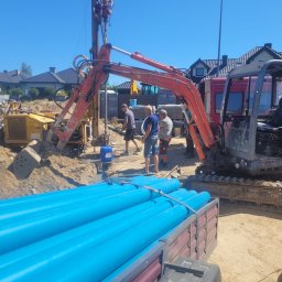 WODAR CENTRUM WIERTNICTWA SPÓŁKA Z OGRANICZONĄ ODPOWIEDZIALNOŚCIĄ - Wiertnica na podwoziu gąsienicowym z ekipą w trakcie wiercenia otworu w ziemi, przygotowane niebieskie rury PCV do wpuszczenia w otwór, w tle zabudowa jednorodzinna.