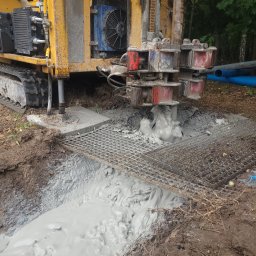 WODAR CENTRUM WIERTNICTWA SPÓŁKA Z OGRANICZONĄ ODPOWIEDZIALNOŚCIĄ - Żółta wiertnica na gąsienicach w trakcie wiercenia otworu w ziemi, z widocznym szlamem i metalową kratą zabezpieczającą wykop.