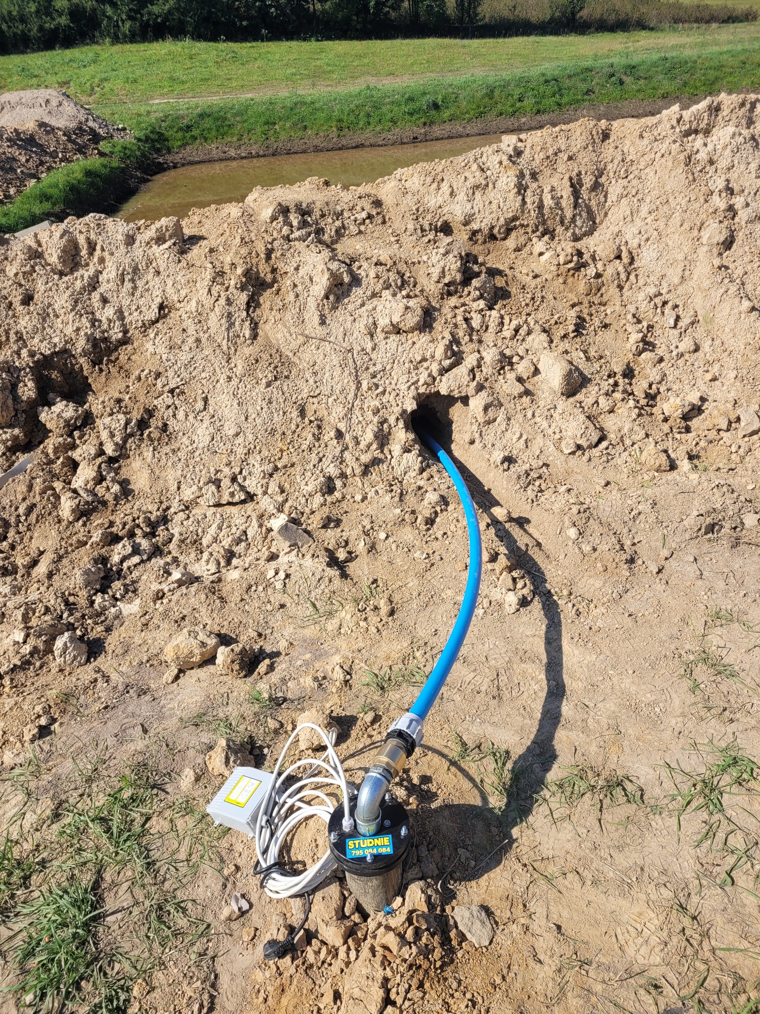 Wiercenie w ziemi z wystającą niebieską rurą, z widoczną pompą i przewodami, na tle wykopu i zieleni.