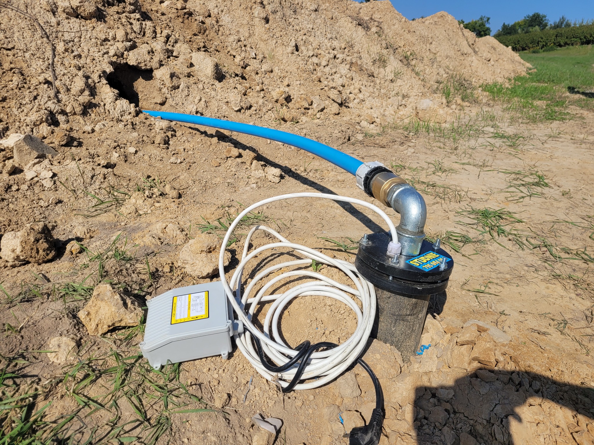Instalacja studni na piaszczystym terenie: niebieska rura wystaje z wykopu, podłączona do metalowego złącza i czarnej obudowy z naklejką, obok szare pudełko sterujące i zwinięty kabel zasilający.