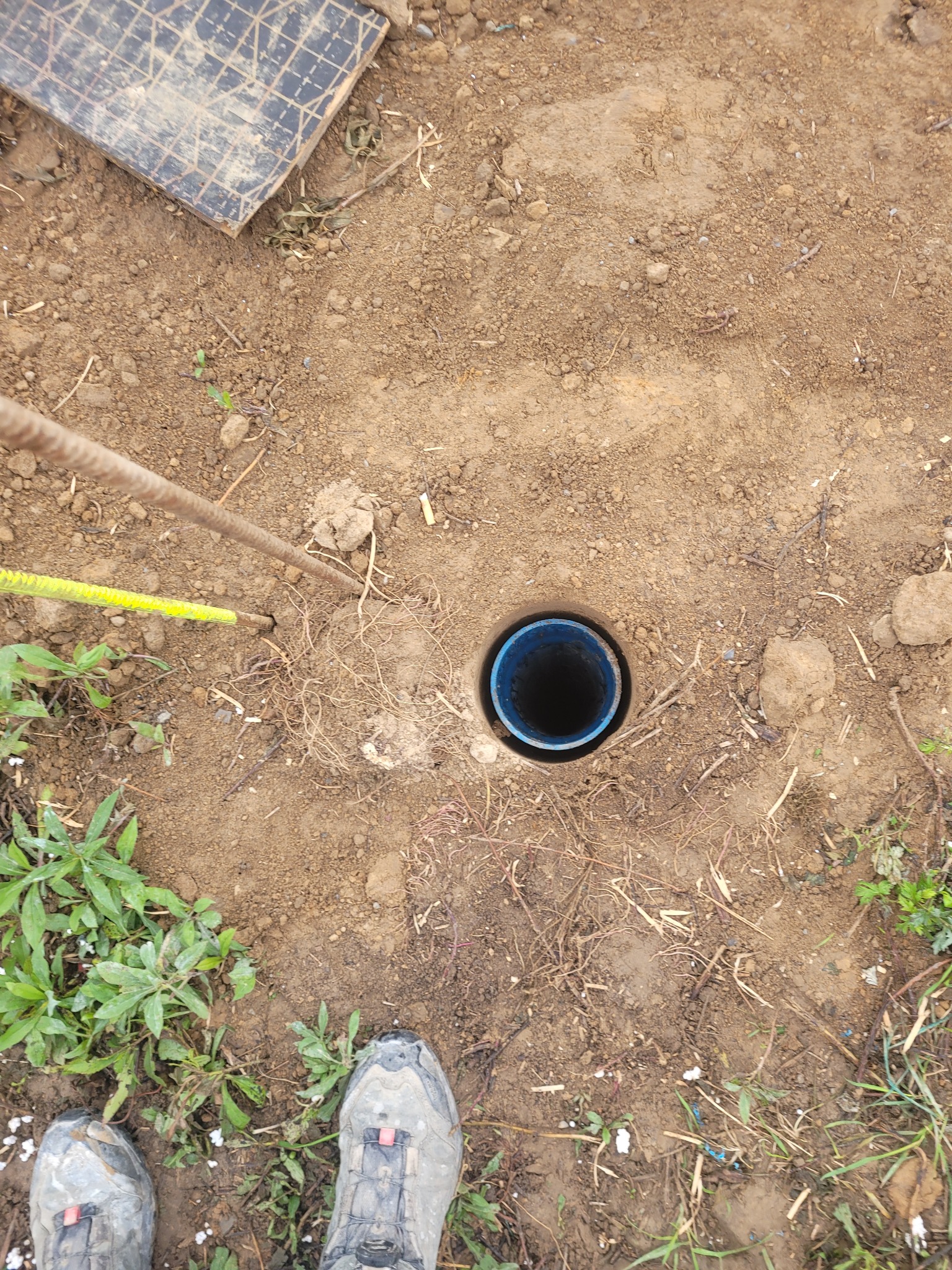 Wykopany otwór w ziemi z niebieską rurą w środku, obok widoczne narzędzia pomiarowe, buty robocze i roślinność.
