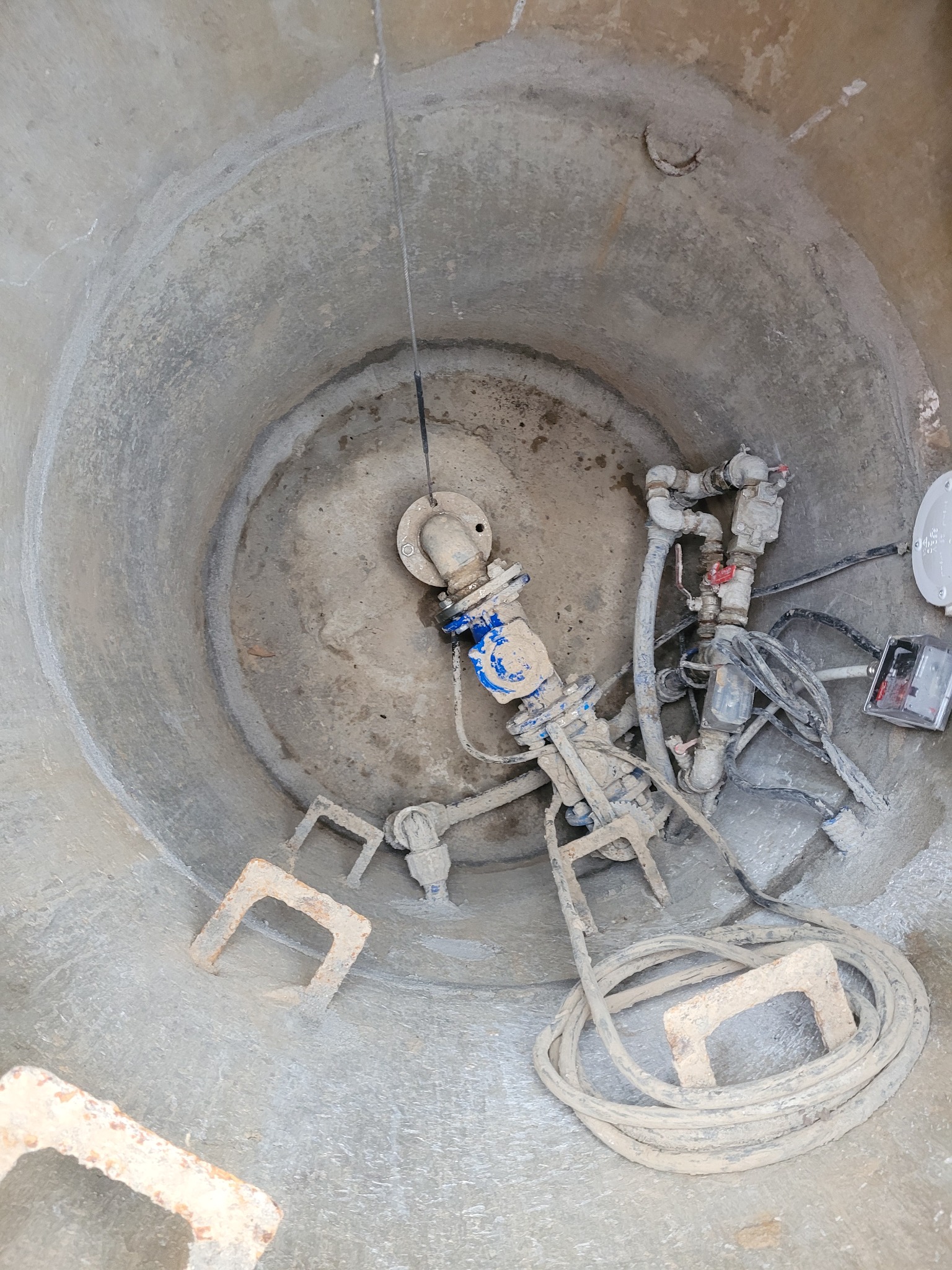 Wnętrze betonowej studni kręgowej z zamontowaną armaturą wodną, widoczne drabinki ze stali nierdzewnej i zwój kabla zasilającego.