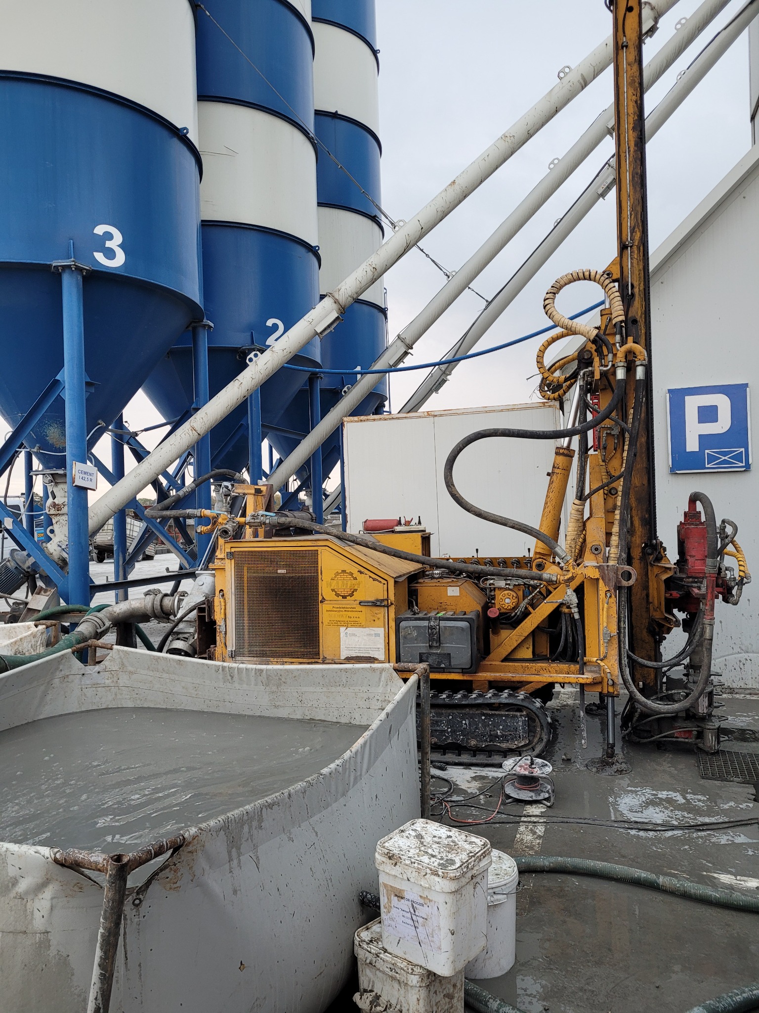 Żółta wiertnica gąsienicowa w trakcie pracy, obok zbiornik z szarą cieczą, w tle silosy na cement i znak parkingu.