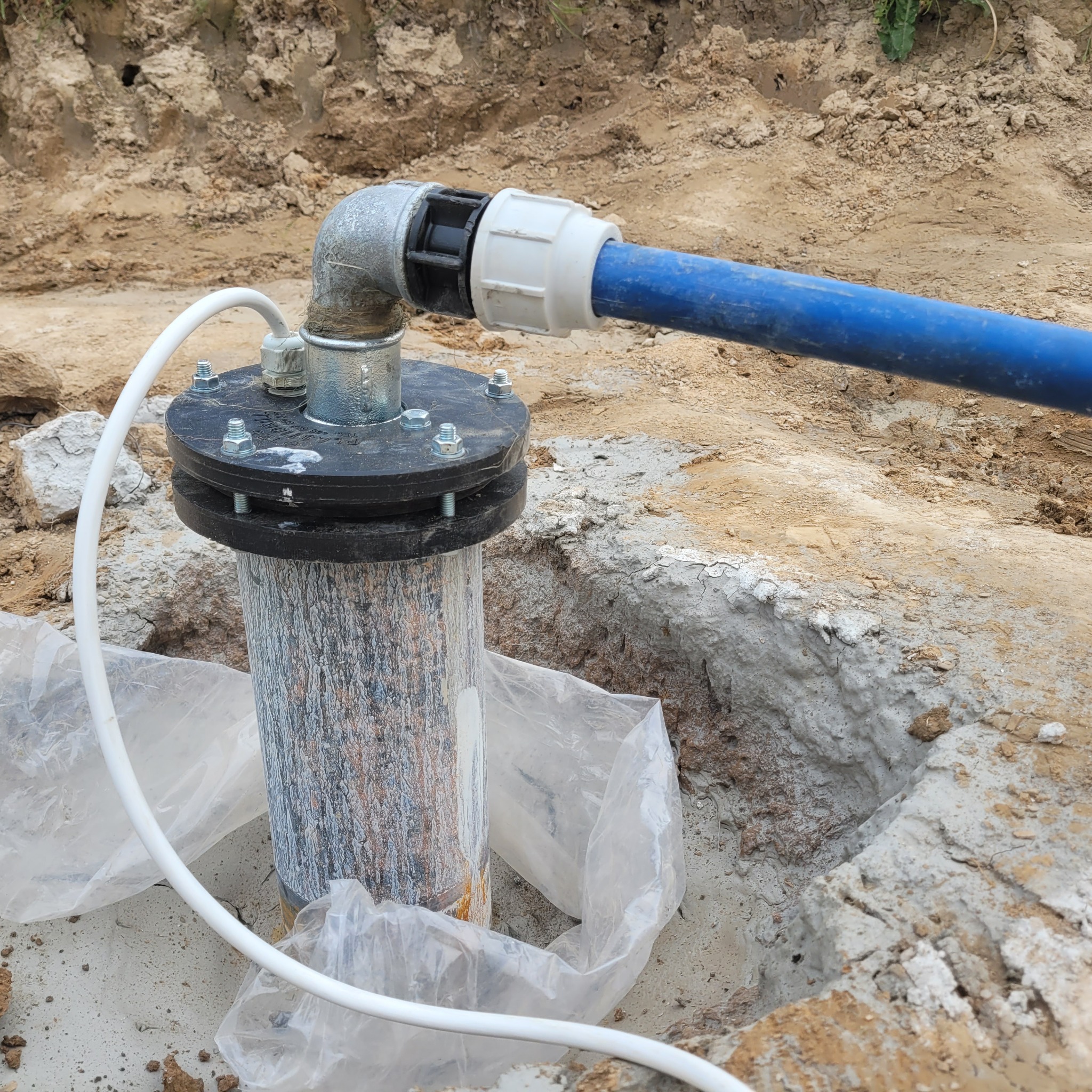 Zakończona instalacja studni z widoczną głowicą, rurą doprowadzającą wodę i kablem zasilającym pompę, umieszczona w wykopie ziemnym.