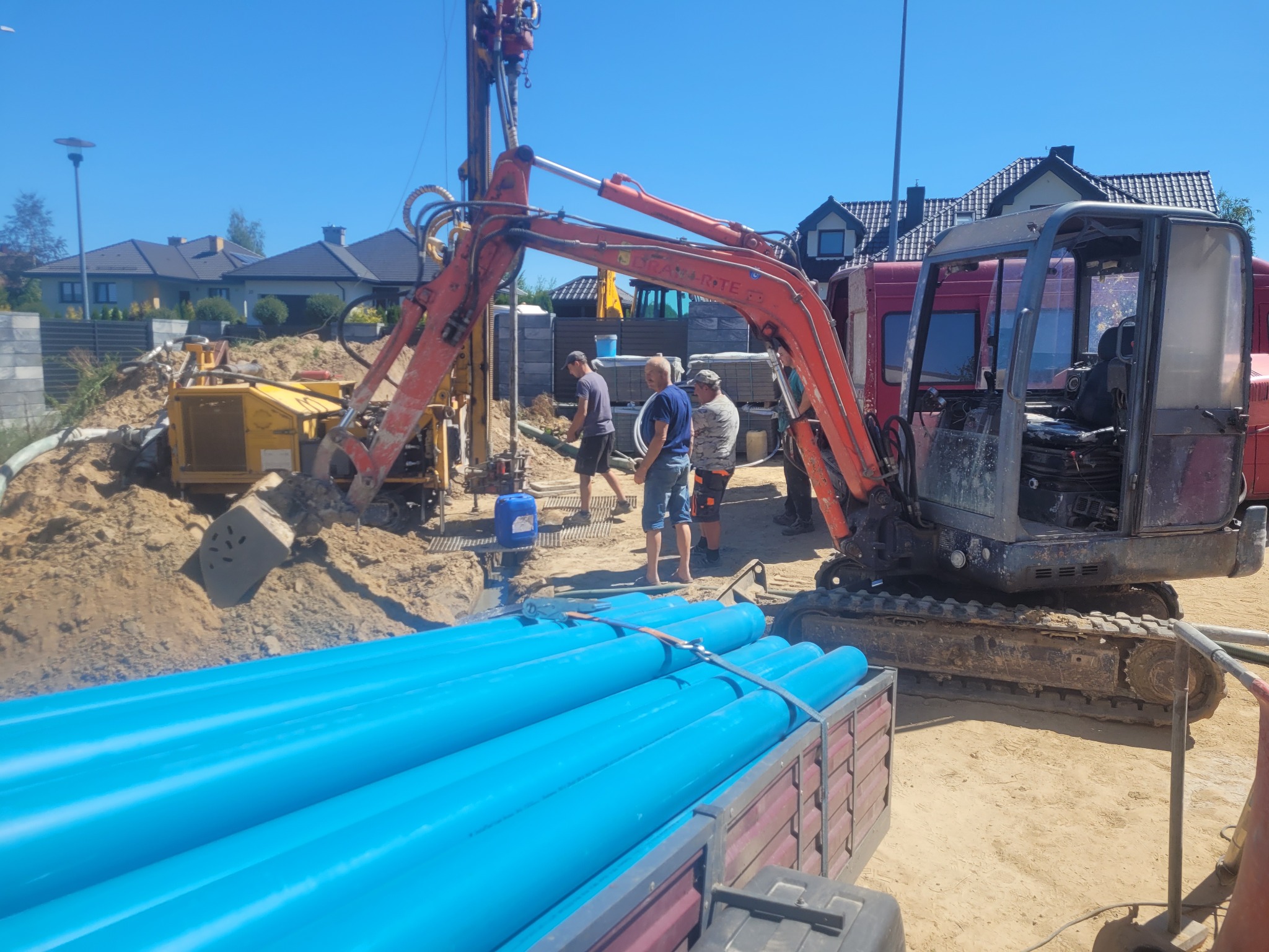 Wiertnica na podwoziu gąsienicowym z ekipą w trakcie wiercenia otworu w ziemi, przygotowane niebieskie rury PCV do wpuszczenia w otwór, w tle zabudowa jednorodzinna.