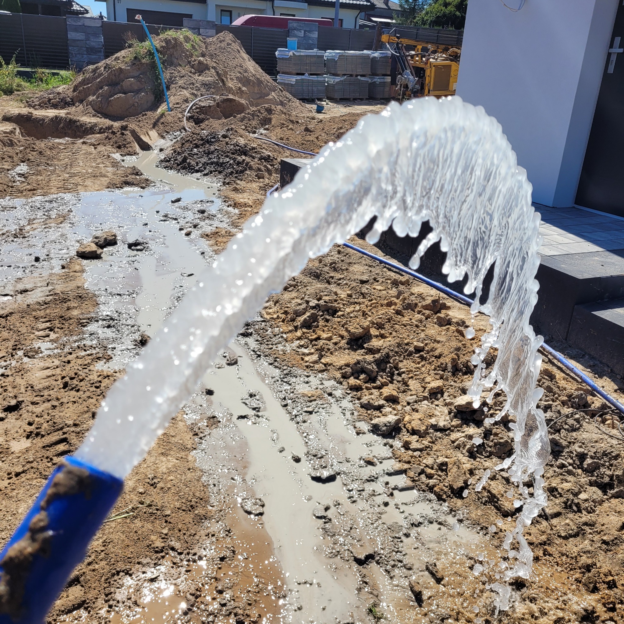 Woda tryskająca z niebieskiego węża na placu budowy, w tle wykopana ziemia, palety z kostką brukową i sprzęt budowlany.