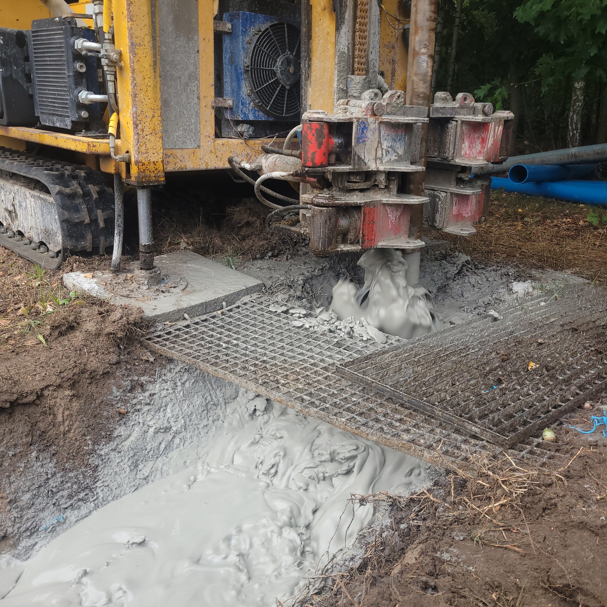 Żółta wiertnica na gąsienicach w trakcie wiercenia otworu w ziemi, z widocznym szlamem i metalową kratą zabezpieczającą wykop.