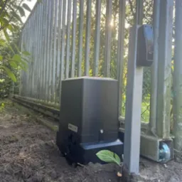 Zamontowany napęd bramy przesuwnej na ziemi, widoczna metalowa brama z pionowych prętów. Słońce prześwituje przez pręty bramy. Nowoczesny design.