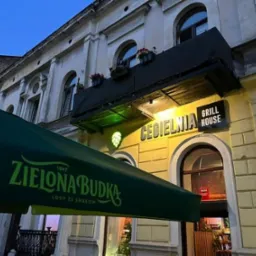Oświetlony budynek Cegielni Grill House nocą, z zielonym baldachimem ZIELONA BUDKA. Widoczne detale architektoniczne i oświetlenie zewnętrzne.