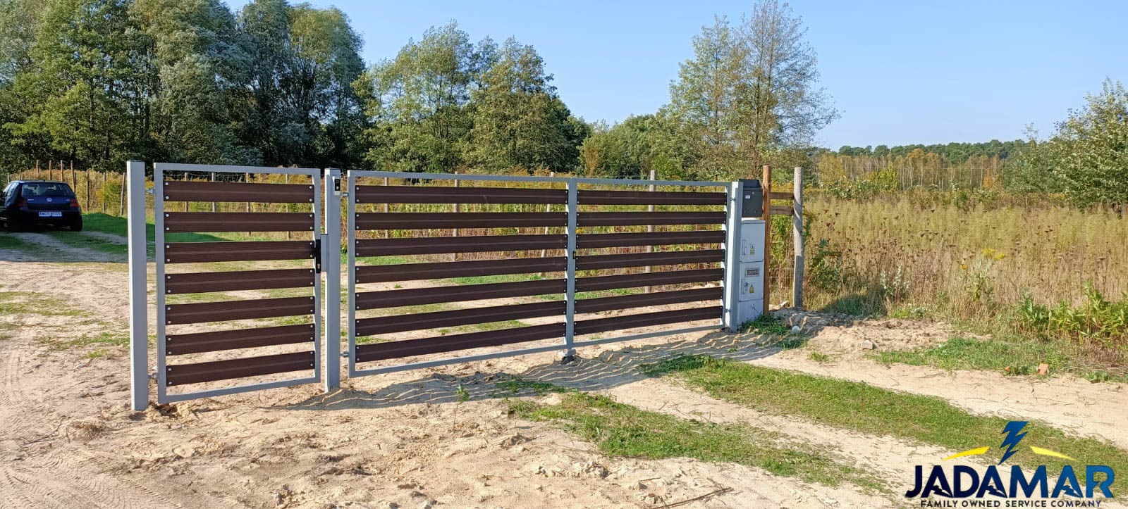 Nowoczesna brama posesyjna z poziomych, brązowych paneli kompozytowych i szarą, metalową ramą, skrzydło bramy otwarte, w tle zaparkowany samochód i zieleń.
