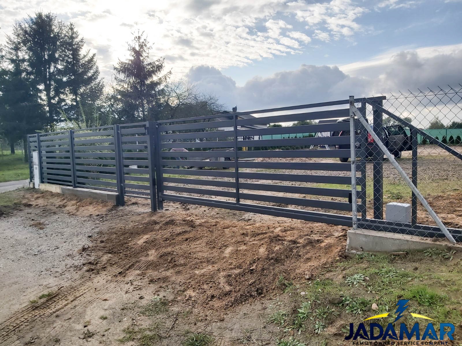 Nowoczesna, przesuwna brama w kolorze grafitowym z poziomymi panelami, zamontowana na betonowej podstawie, widoczna w trakcie instalacji, z częściowo naruszonym terenem wokół.