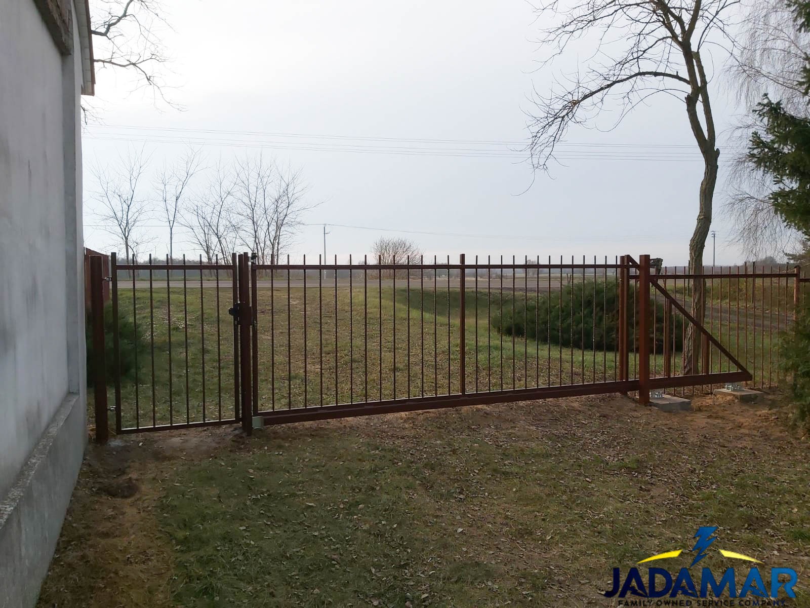 Przesuwna, metalowa brama wjazdowa z pionowymi prętami, pomalowana na brązowo, z widocznym zamkiem, na tle zielonego trawnika i drzew bez liści, z szarym niebem w tle.