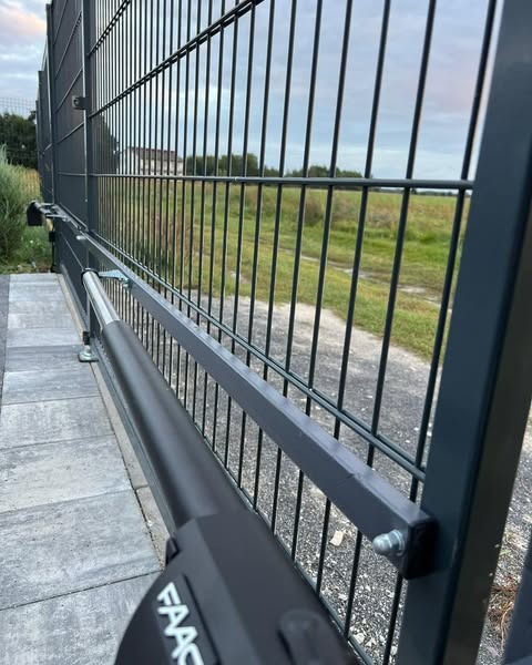 Szara brama panelowa z zamontowanym siłownikiem FAAC, widok z bliska na mechanizm automatyki. Trawa i zabudowania w tle.