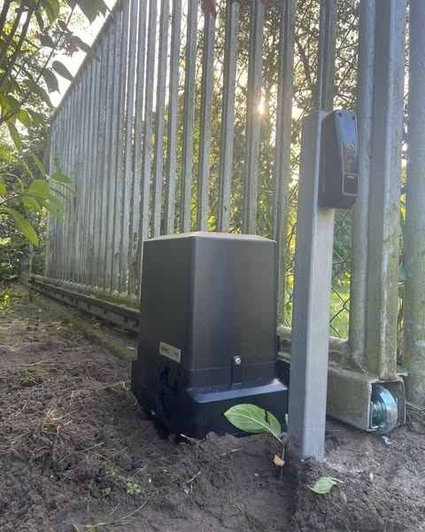 Zamontowany napęd bramy przesuwnej na ziemi, widoczna metalowa brama z pionowych prętów. Słońce prześwituje przez pręty bramy. Nowoczesny design.