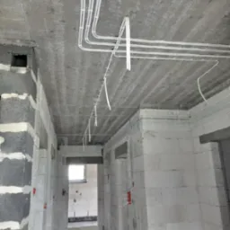 Białe kable elektryczne ułożone na suficie z surowego betonu w budynku w trakcie budowy. Widoczne ściany z białych bloczków i otwory drzwiowe.