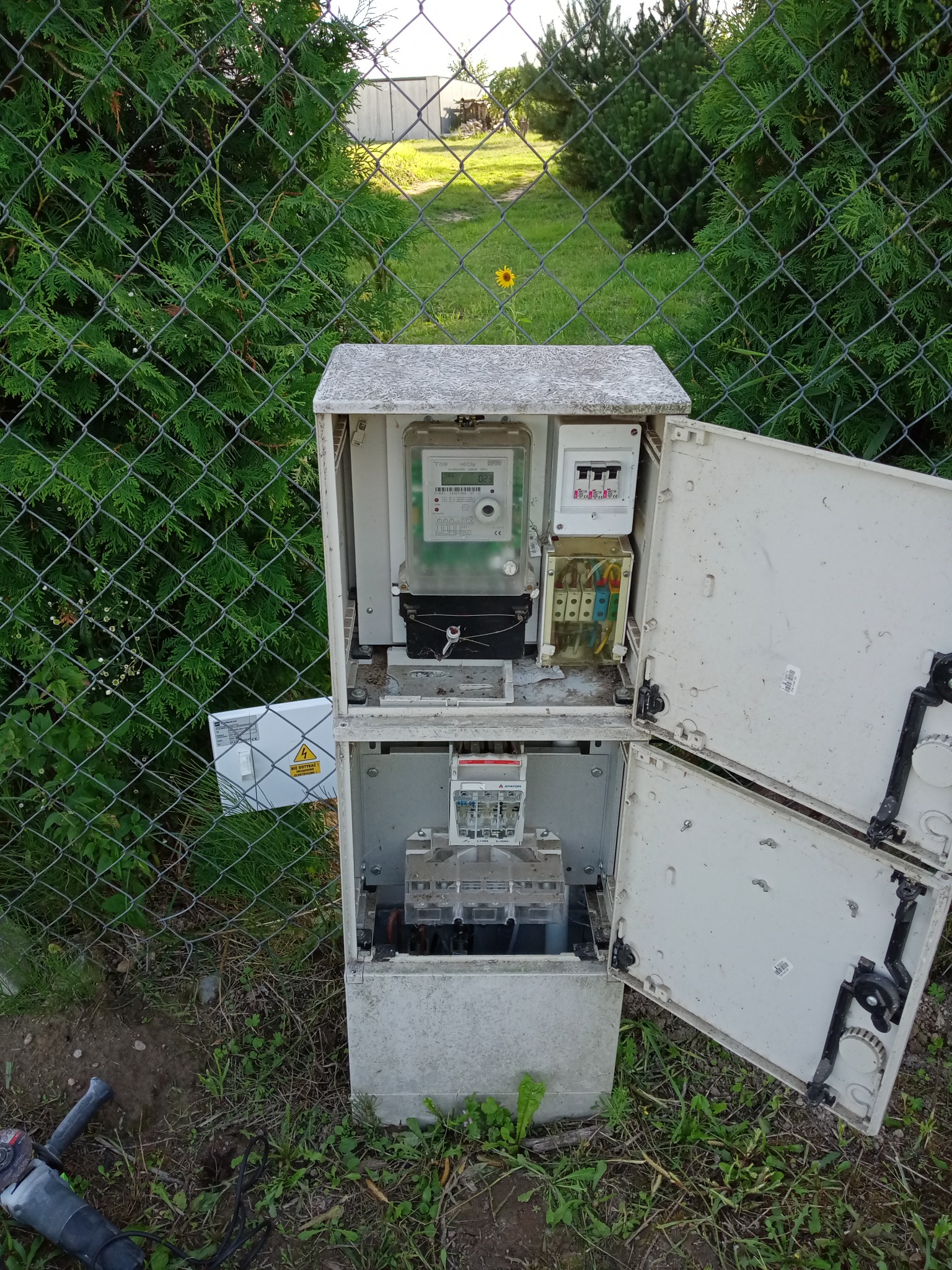 Otwarta skrzynka elektryczna z licznikiem i bezpiecznikami, widoczna na tle zieleni i siatki ogrodzeniowej, z narzędziem szlifierskim obok.