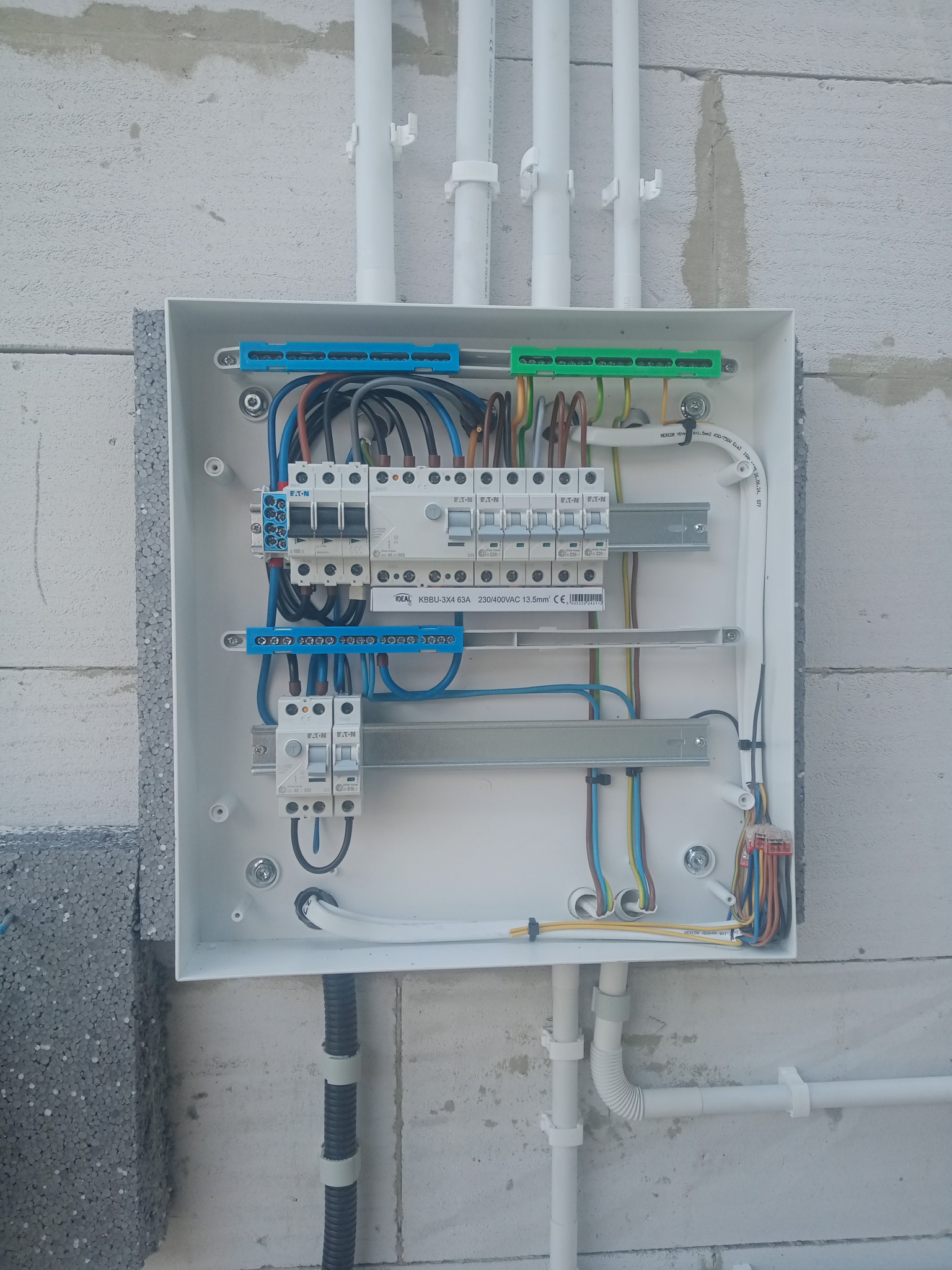 Rozdzielnica elektryczna Eaton zamontowana na ścianie z białego betonu komórkowego. Widoczne kable i przewody. Białe rury PCV przymocowane do ściany.