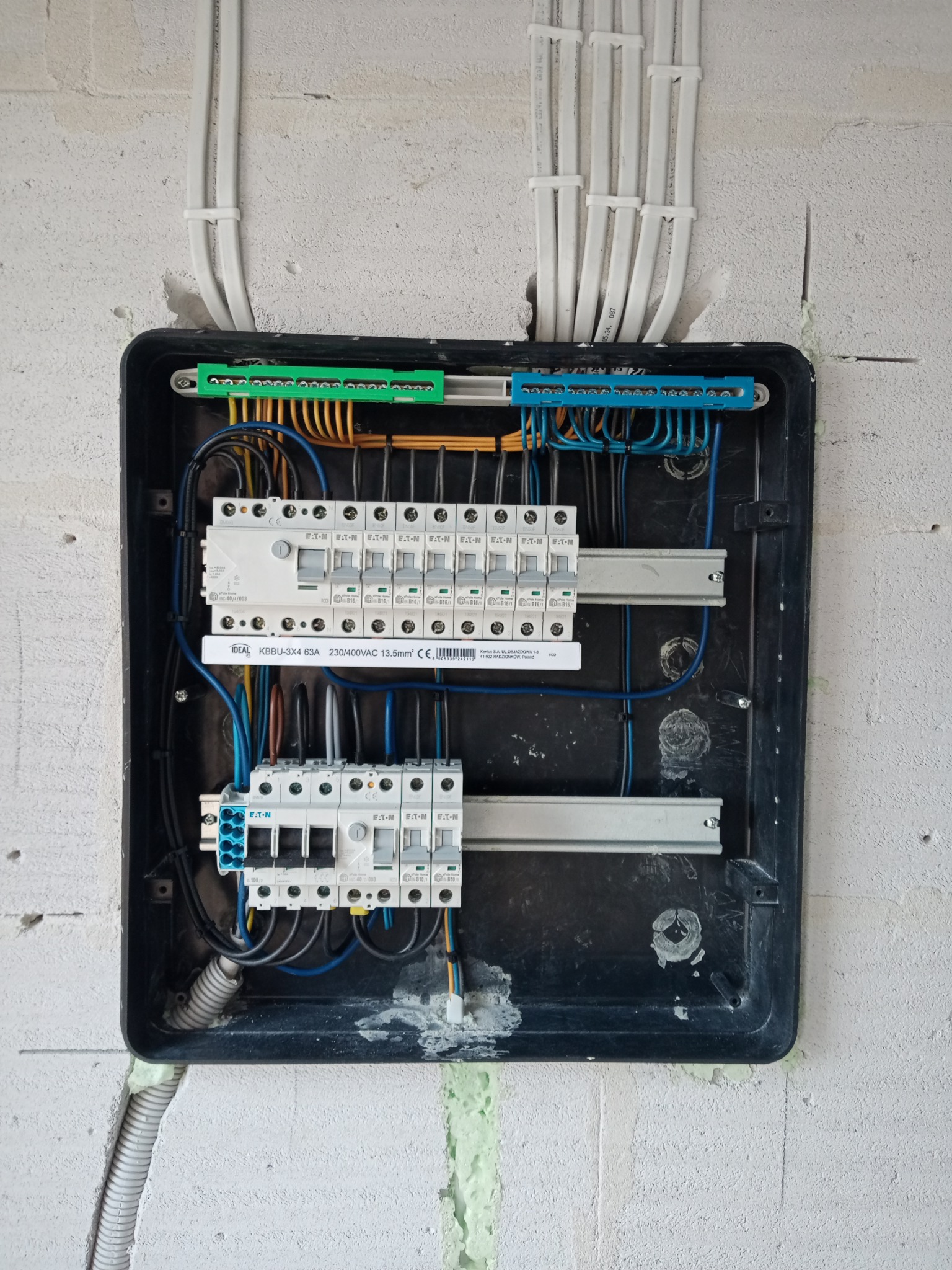 Rozdzielnica elektryczna w trakcie montażu w surowym wnętrzu. Widoczne kable, bezpieczniki i listwy zaciskowe. Ściana z betonu komórkowego.