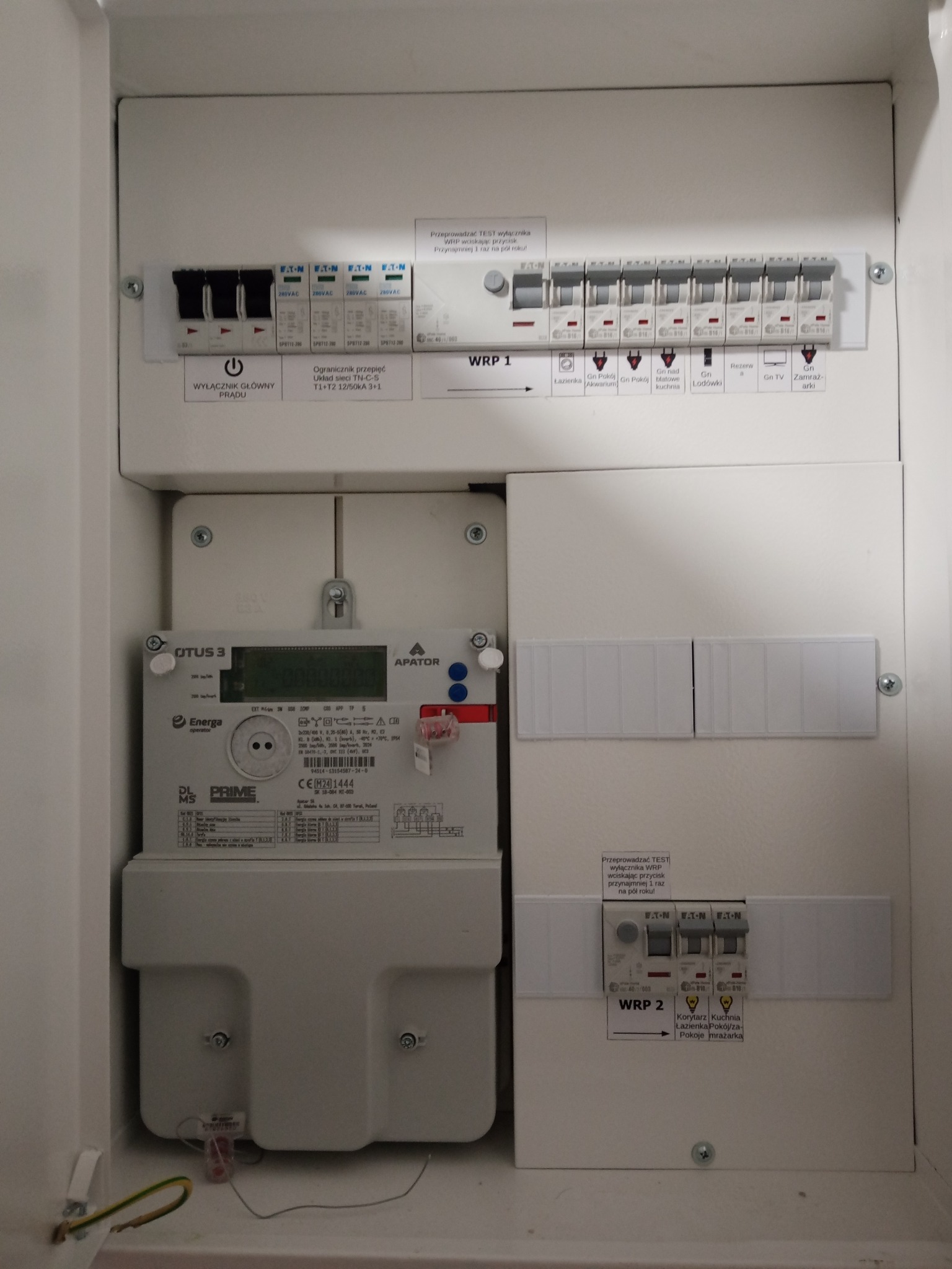 Rozdzielnica elektryczna wnękowa z licznikiem energii, bezpiecznikami i wyłącznikami. Widoczne opisy obwodów i naklejki informacyjne. Całość w białej obudowie.